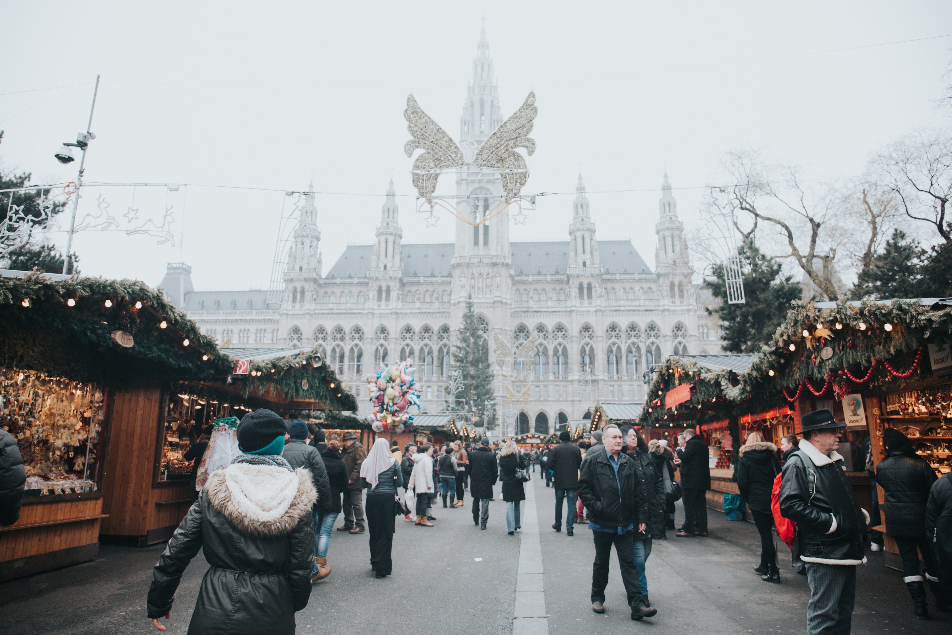 The image shows a bustling Christmas market in Vienna, Austria. There are many people walking...