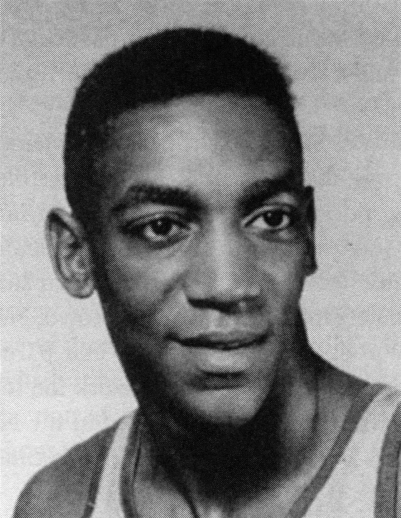 The image shows a black and white photo of a man in a basketball uniform, smiling. He is wearing a...