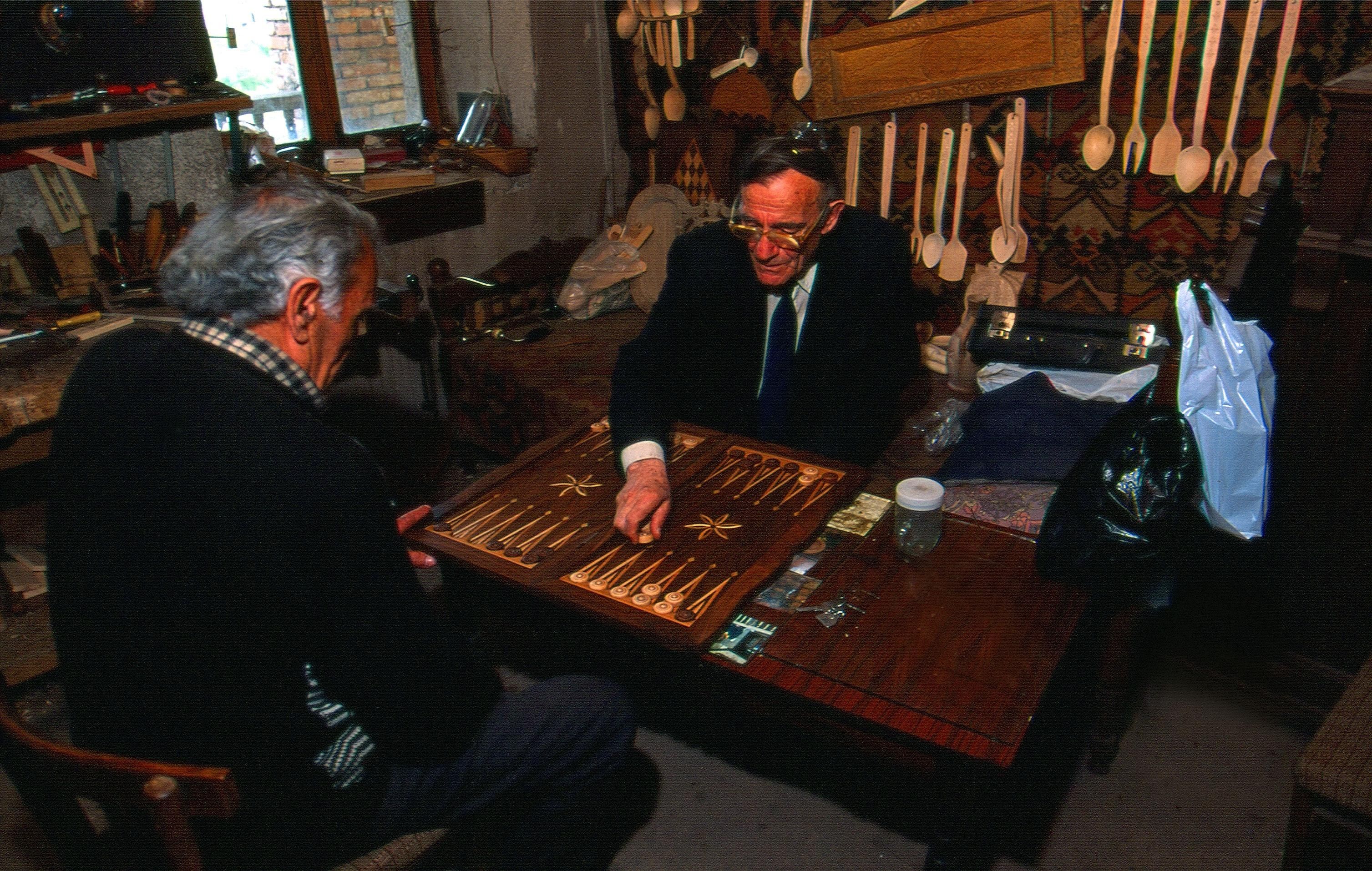 The image shows two men sitting at a table playing a game of backgammon. On the table there are...