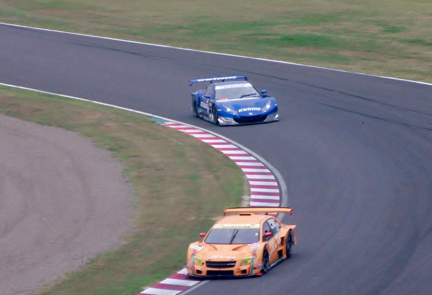 The image shows two sports cars racing on a race track, with one car in orange and the other in...