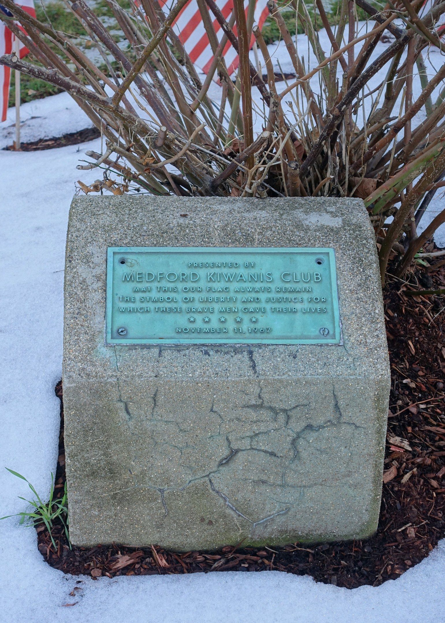The image shows a memorial stone with a plaque on it in the snow, surrounded by plants and a flag...