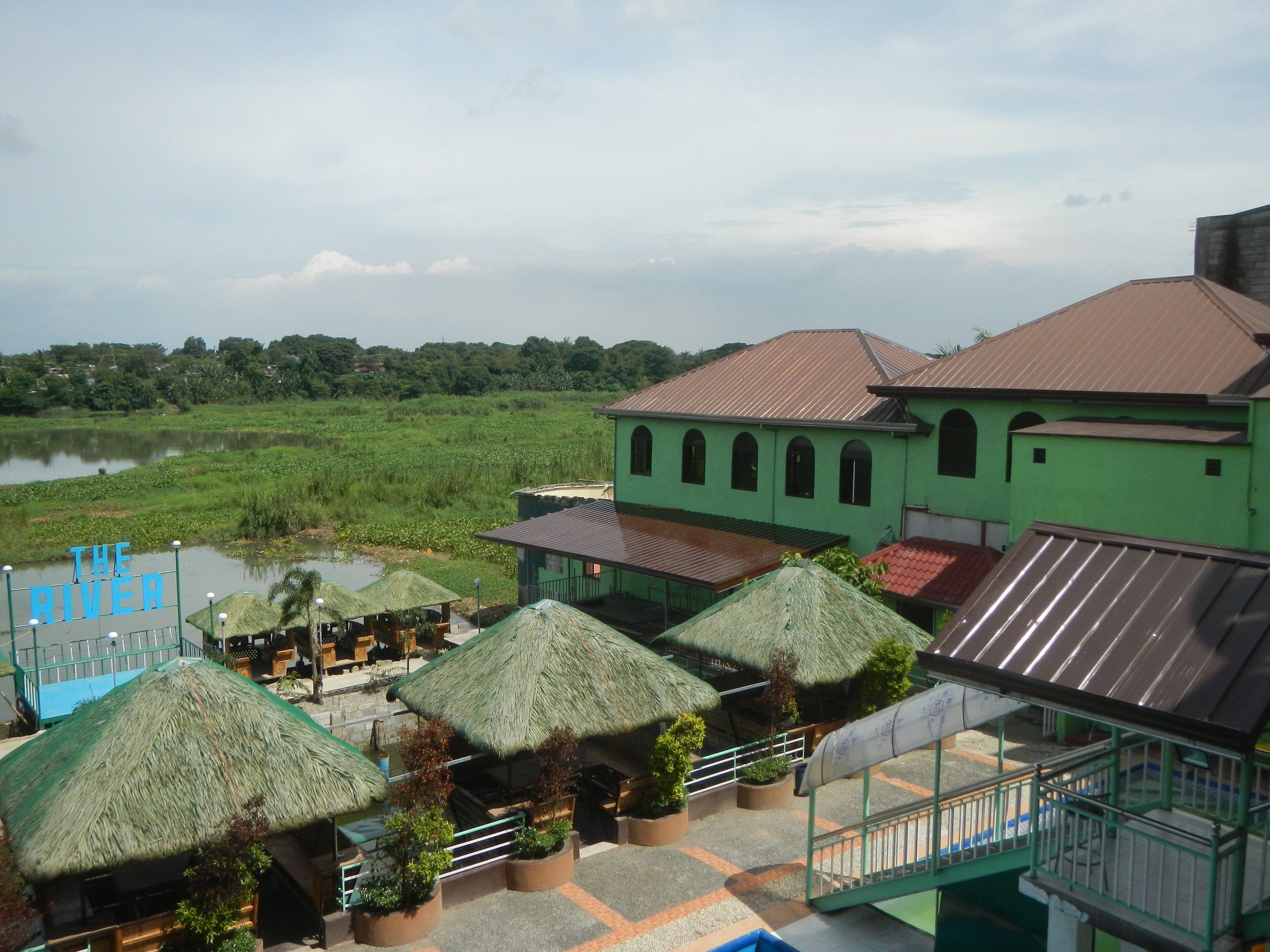 The image shows a view of a resort from the top of a building, with thatched roofs, railings,...