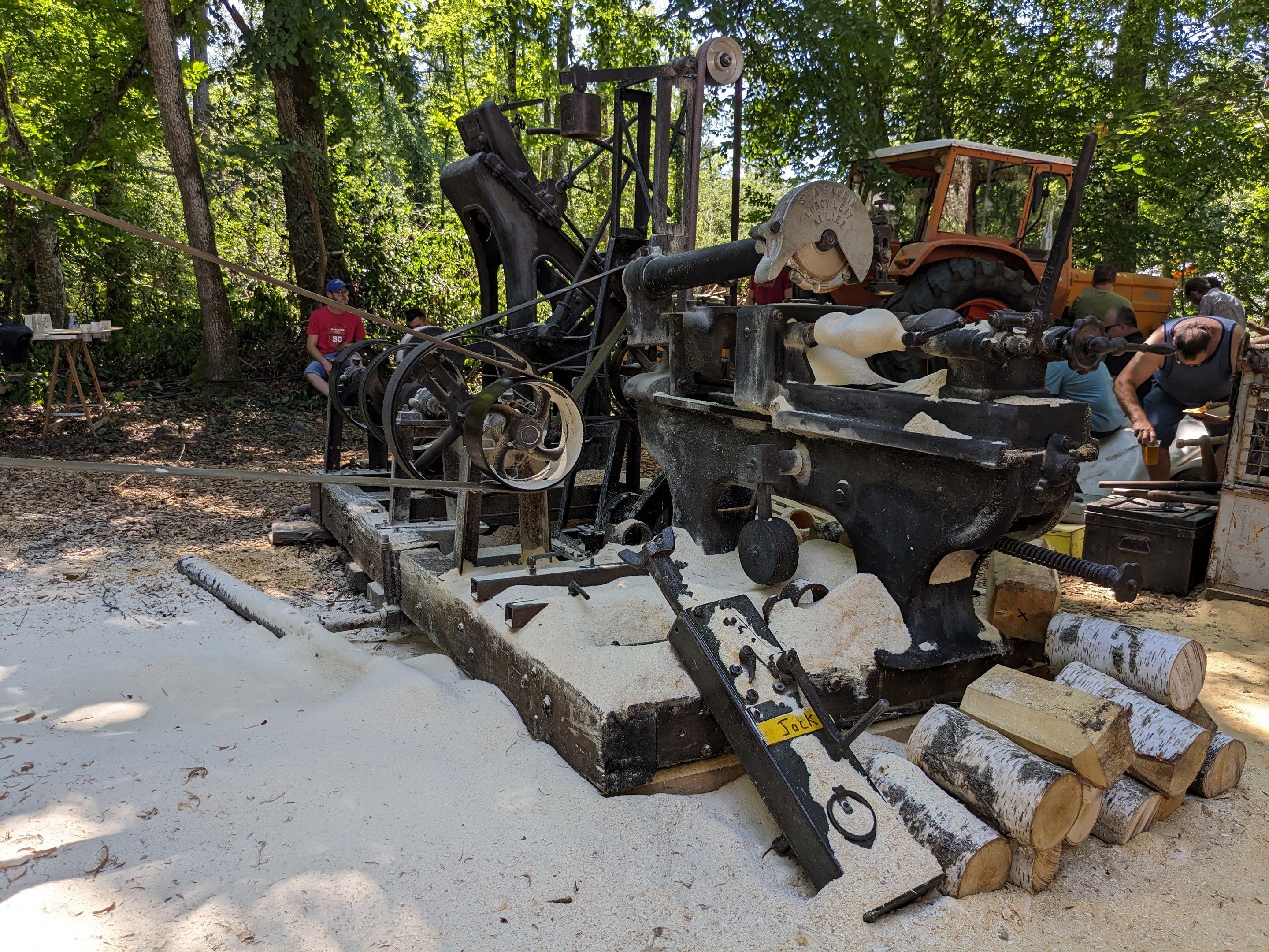 The image shows a group of people working on a machine in the woods. There are wooden logs, a...