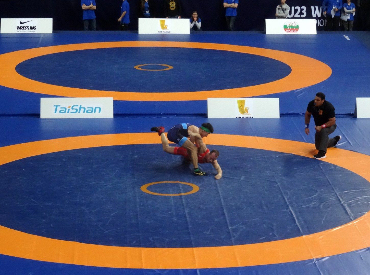 The image shows two wrestlers in a wrestling match on a blue and orange mat, with a few people...