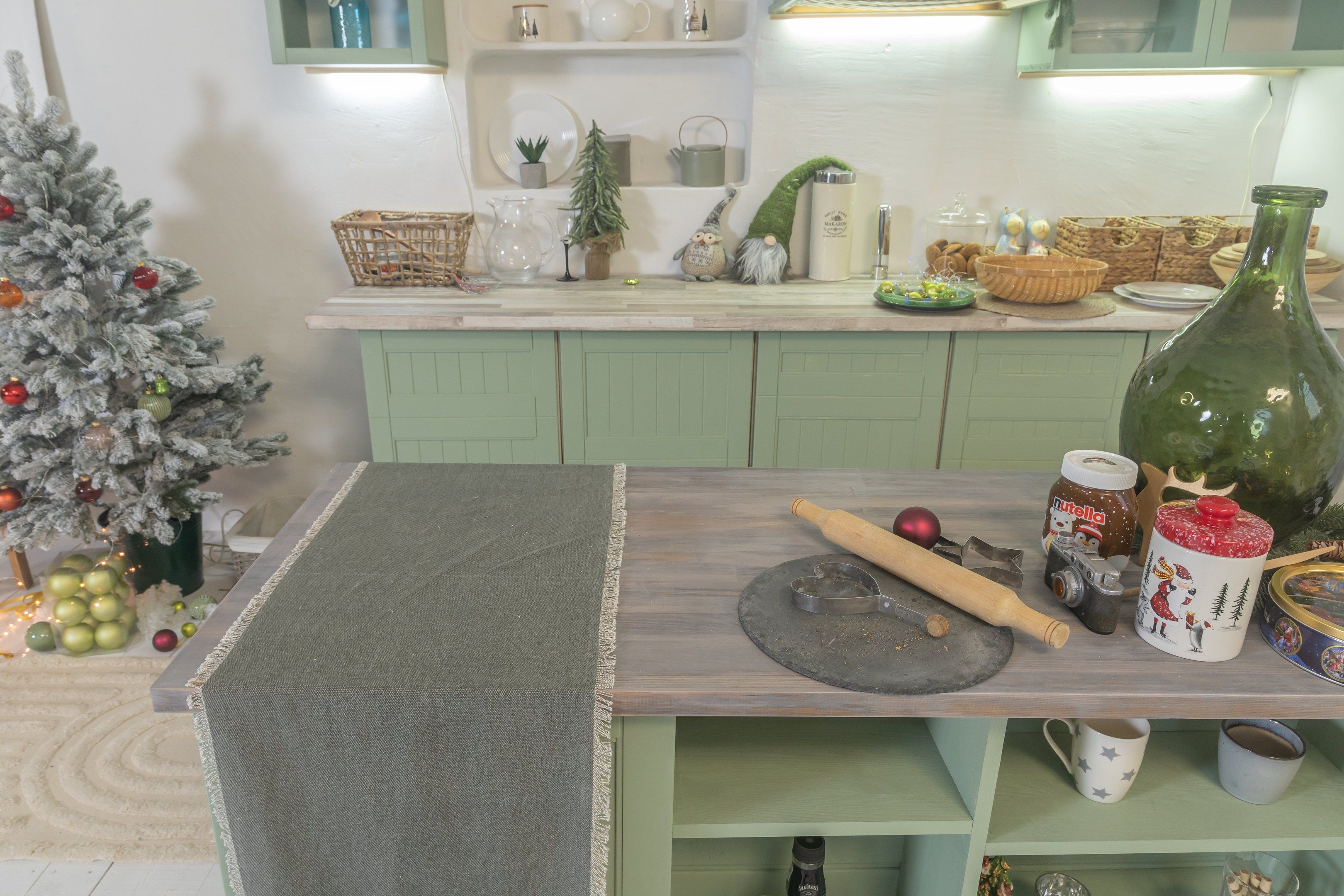 The image shows a kitchen with green cabinets and a Christmas tree in the corner. On the table...