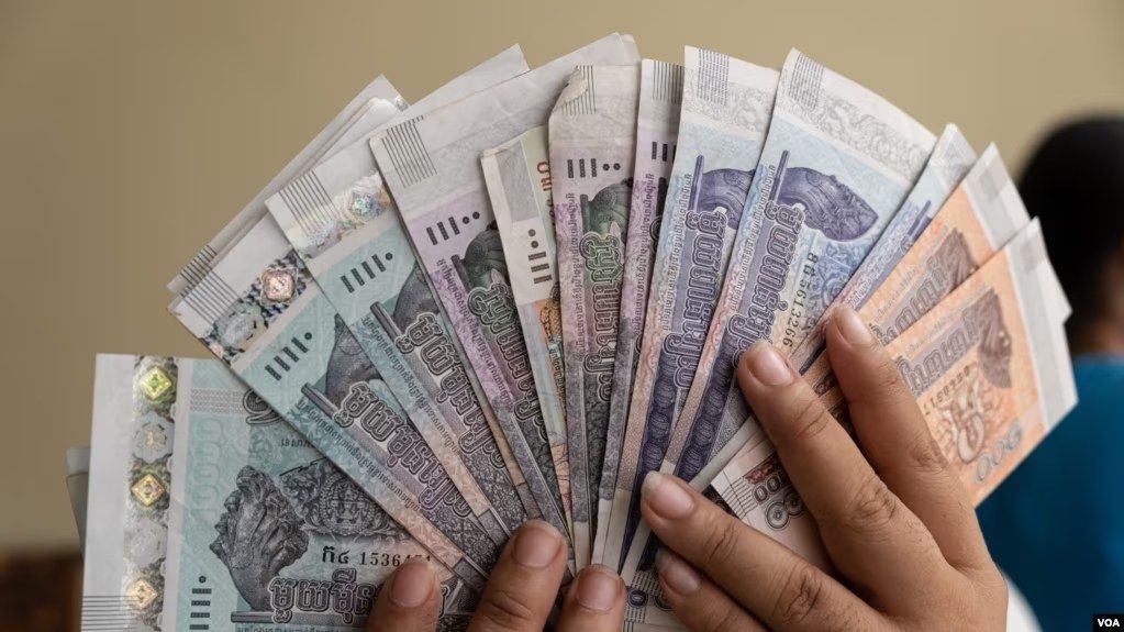 The image shows a person holding a bunch of Thai money in their hands, with a blurred background...