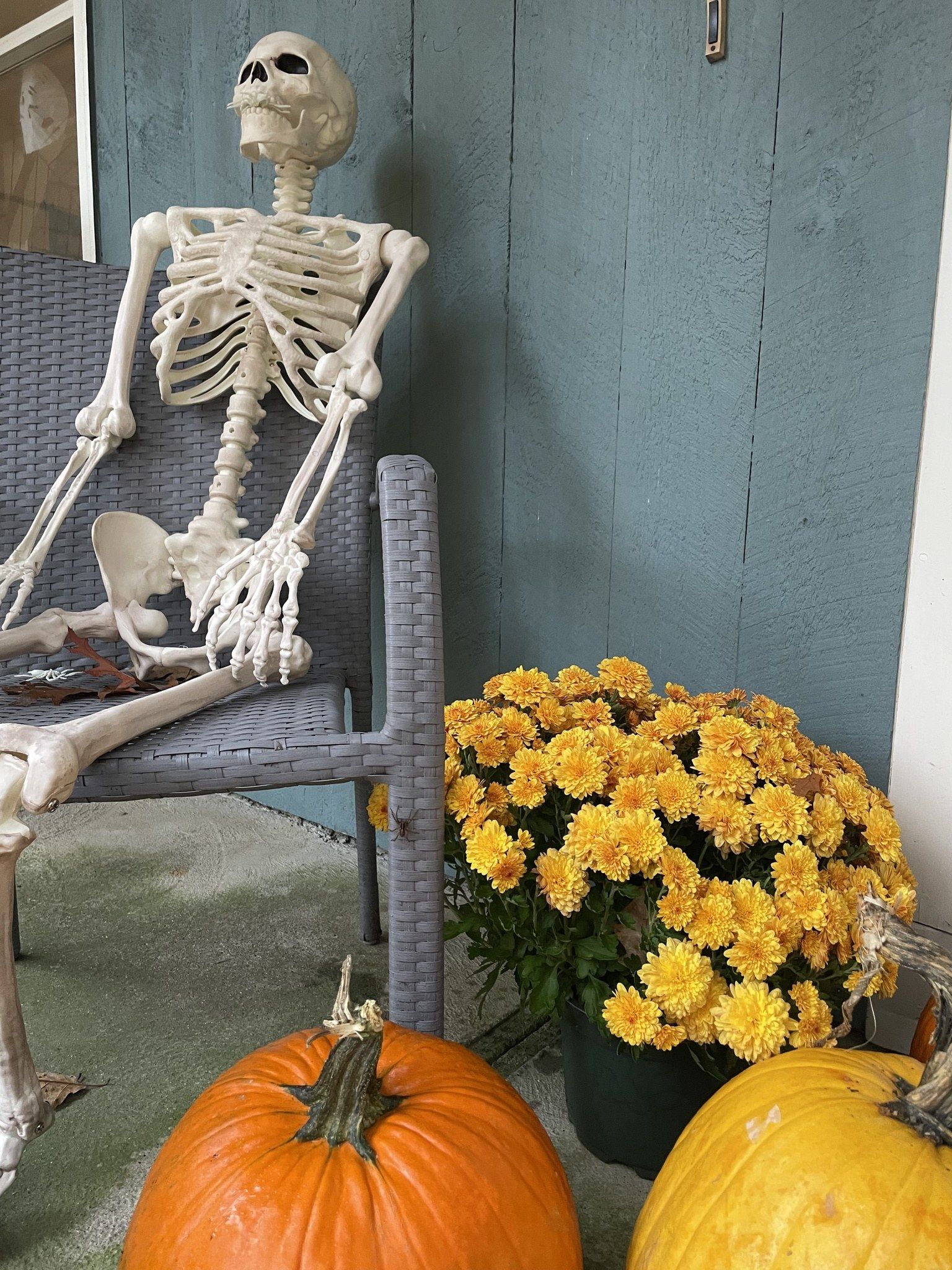 The image shows a skeleton sitting on a chair next to two pumpkins on a porch, surrounded by a...