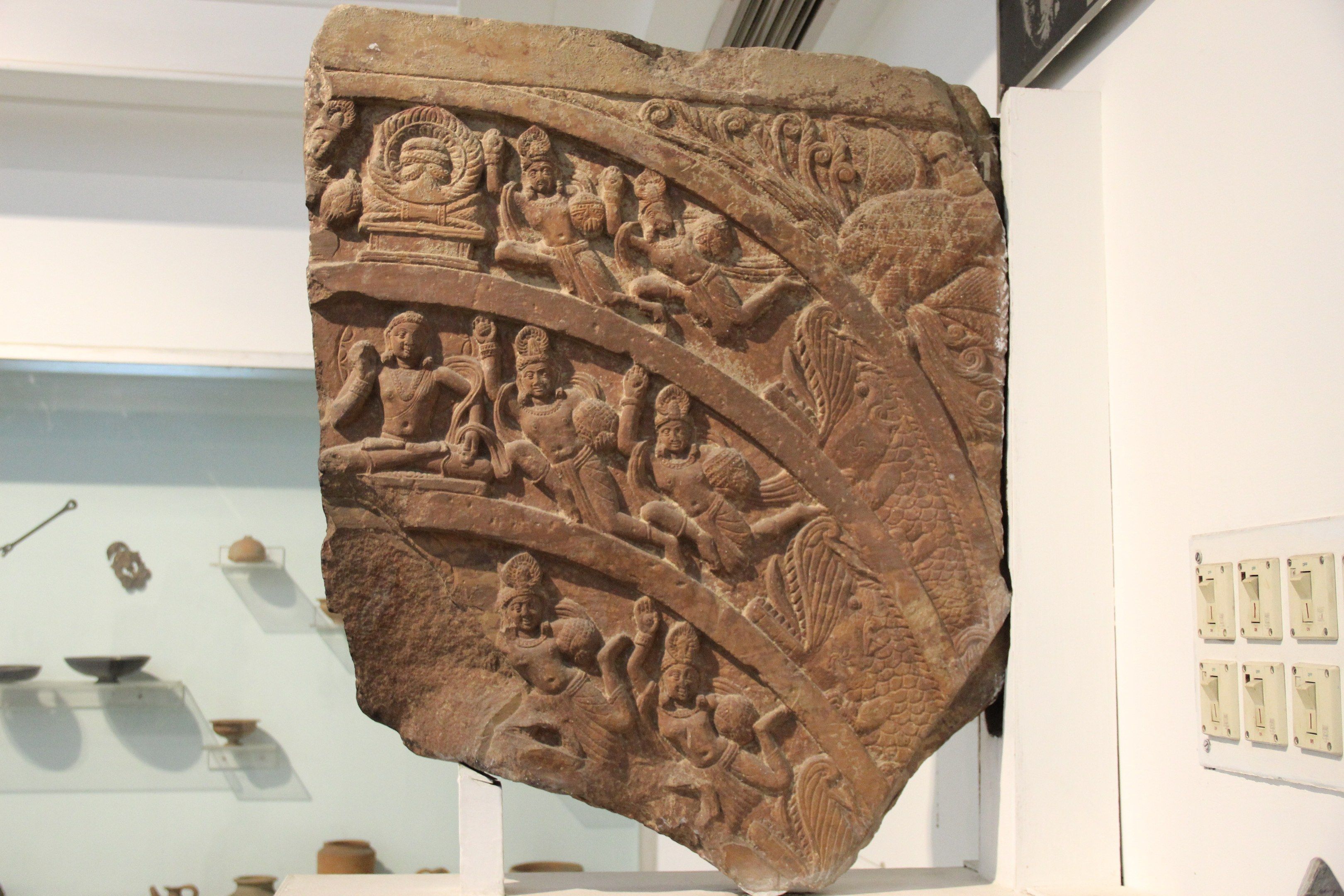 The image shows a large stone relief on display in a museum, with intricate carvings on its...