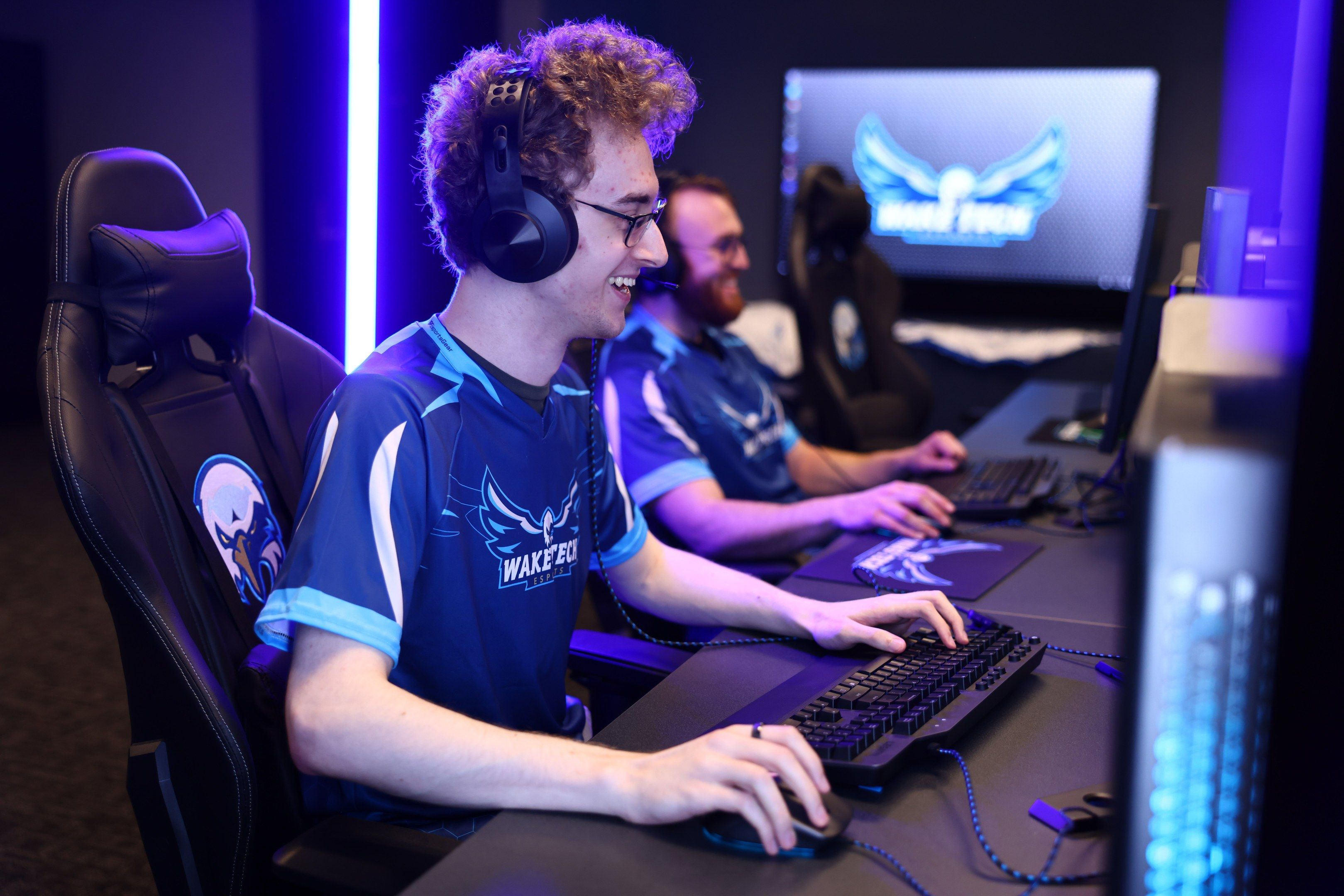 The image shows two men sitting at a desk in front of a computer, wearing headsets and smiling. On...