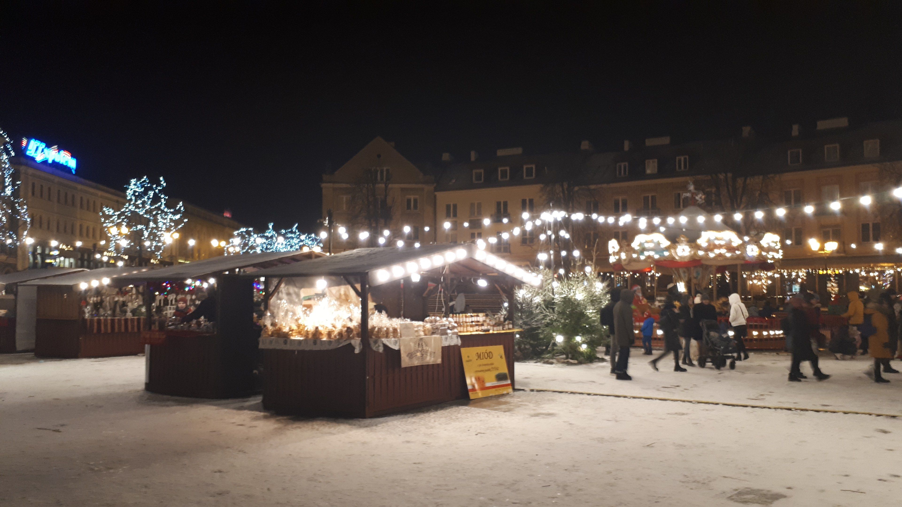 The image shows a bustling Christmas market in the middle of a snowy street at night. We can see a...