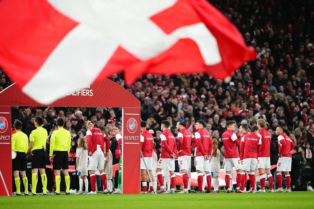 The image shows a group of people standing on top of a soccer field, with a red and white flag in...