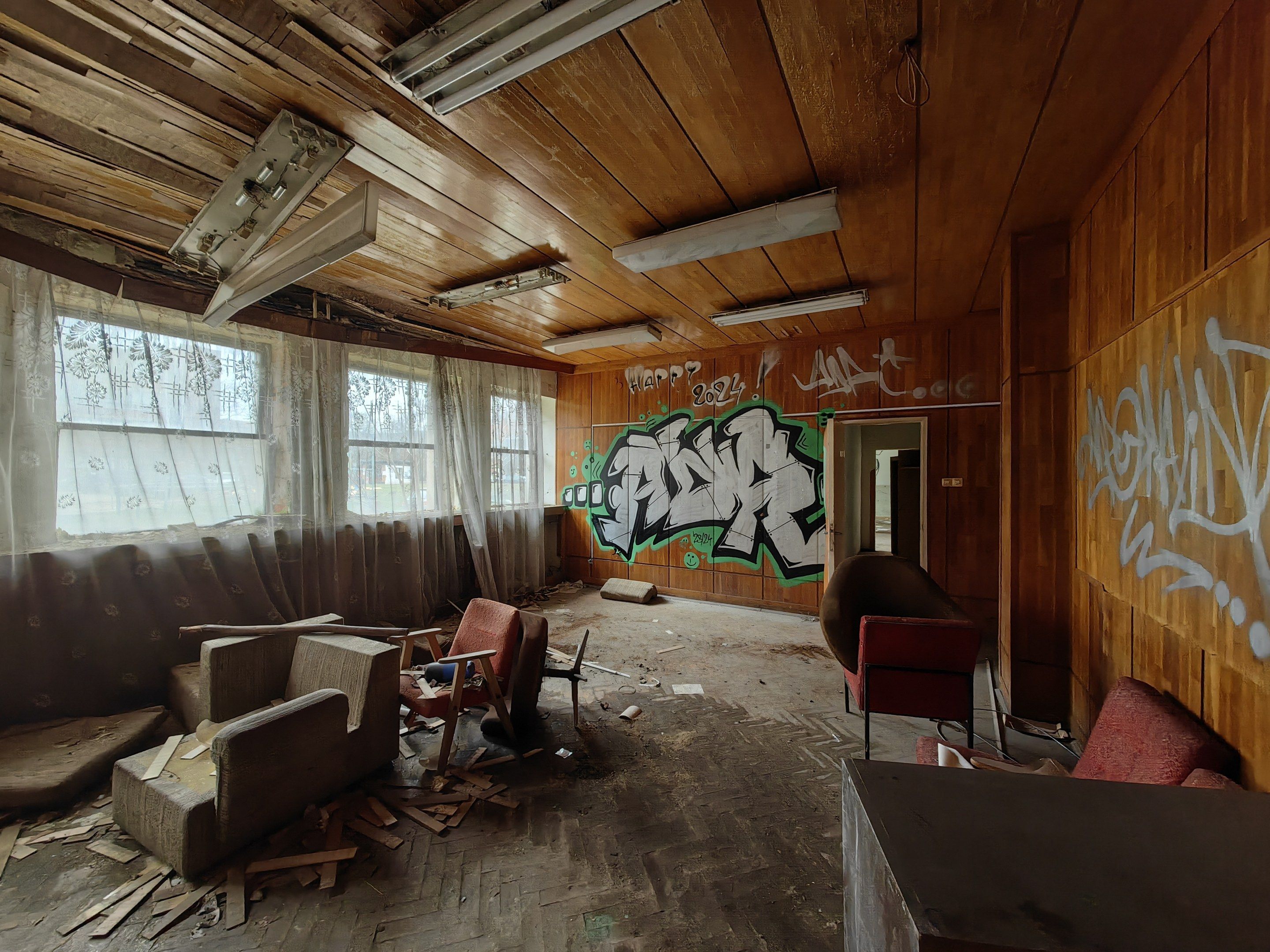 The image shows an empty room with graffiti on the walls, sofas, chairs, wooden objects, windows...