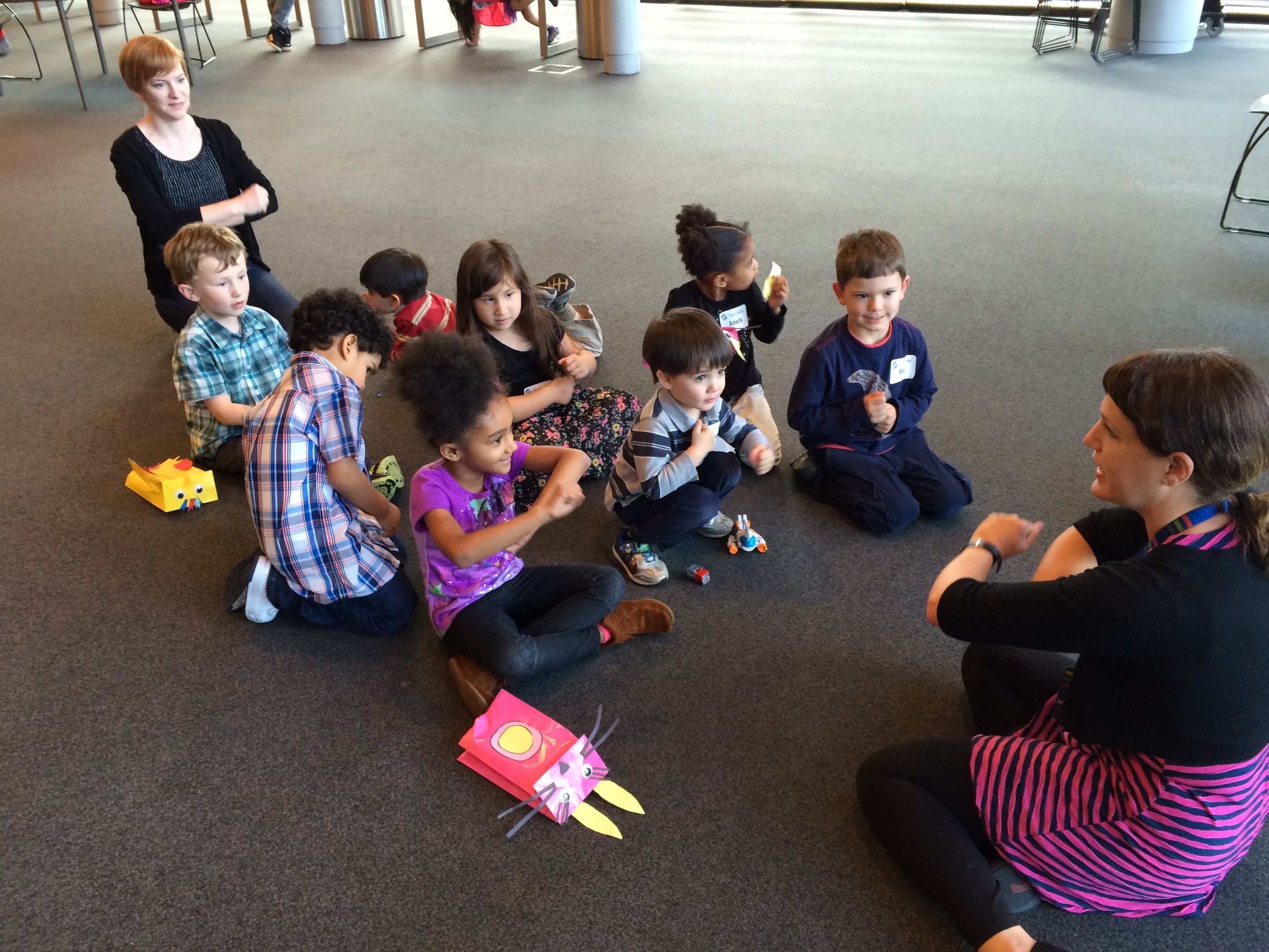 The image shows a group of children sitting on the floor in front of a teacher, surrounded by toys....