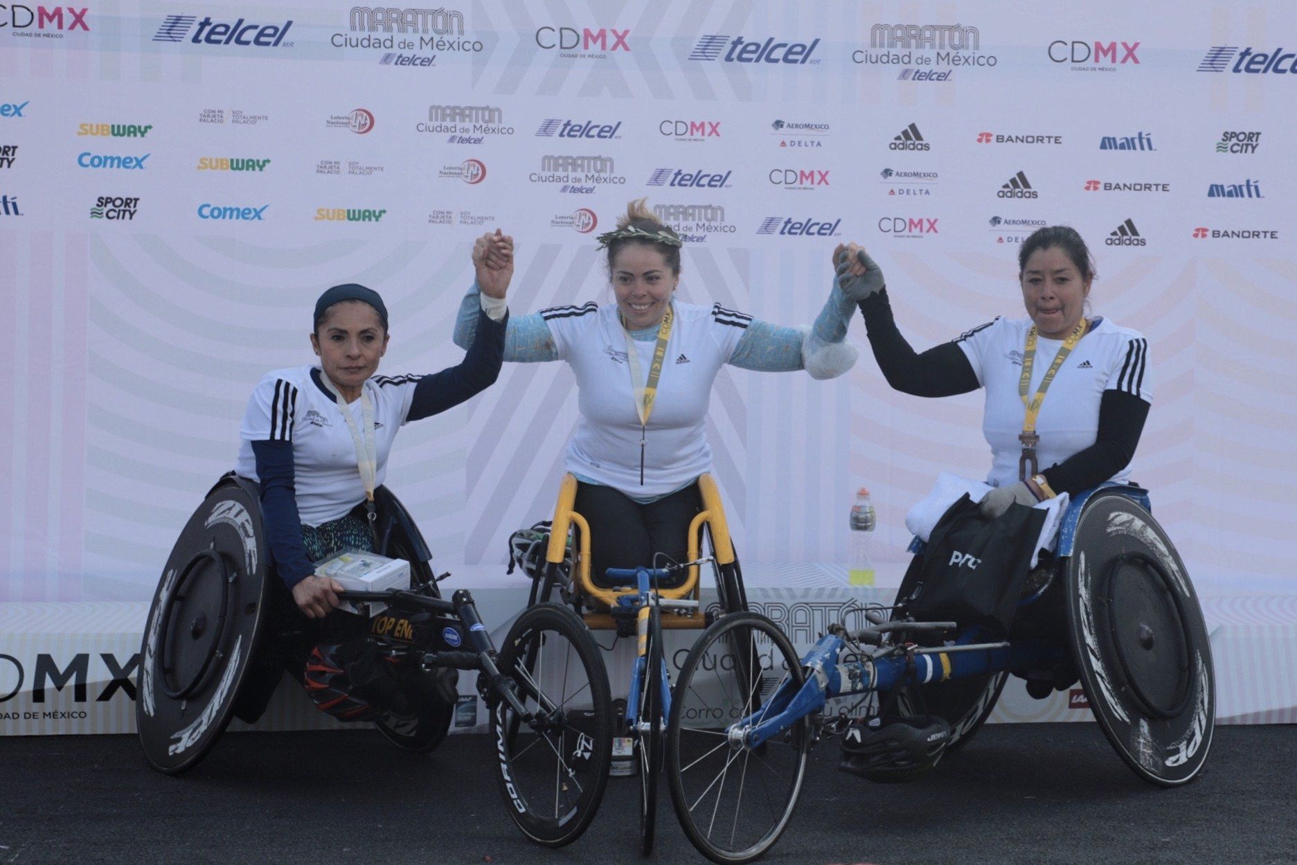 The image shows three women in wheelchairs standing on a podium with their hands in the air,...