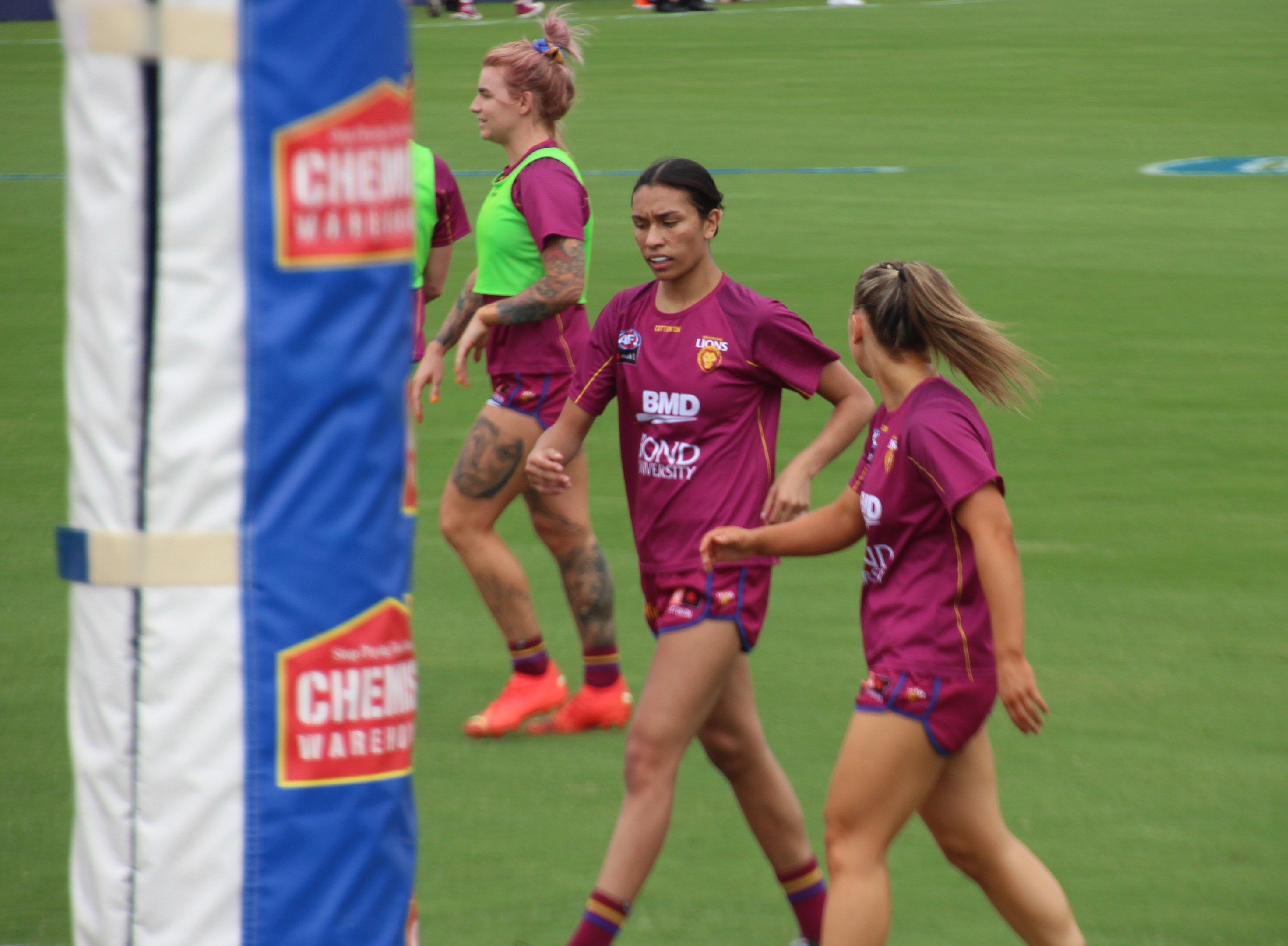 The image shows a group of women walking across a lush green field, with a pole on the left side of...