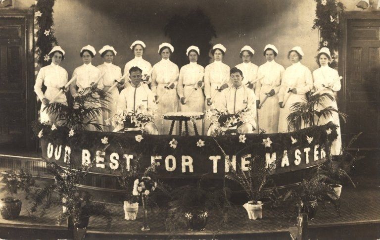 The image shows a group of women standing on top of a stage, wearing white dresses and caps. In the...