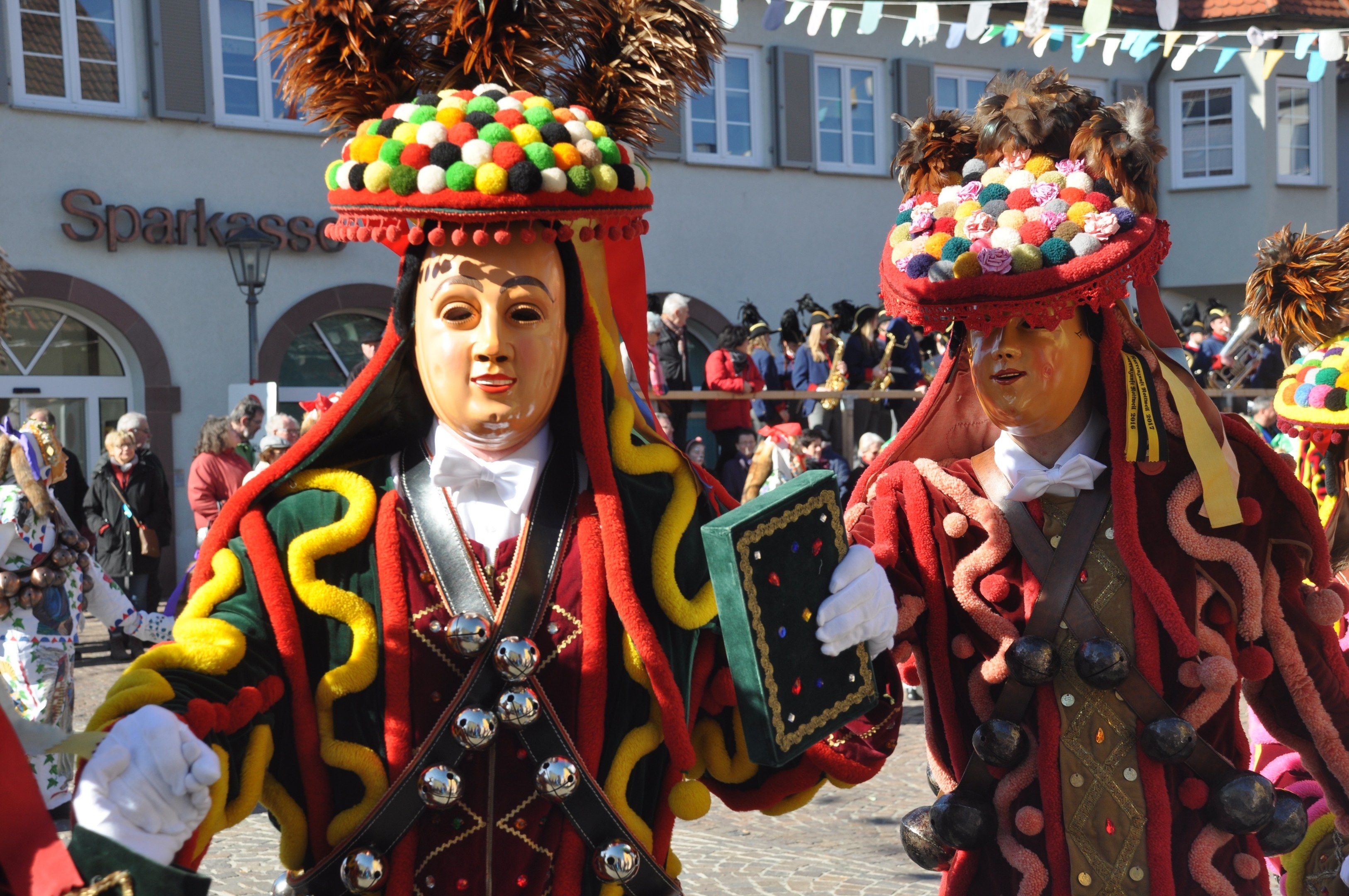 The image shows a group of people in colorful costumes walking down a street, with a building in...