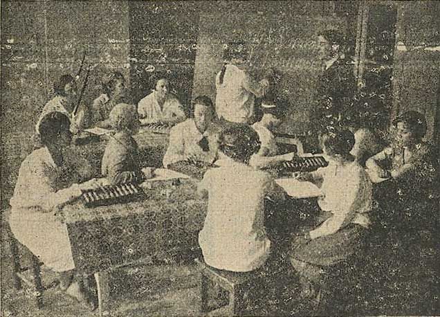 The image shows an old black and white photo of a group of people sitting around a table playing...