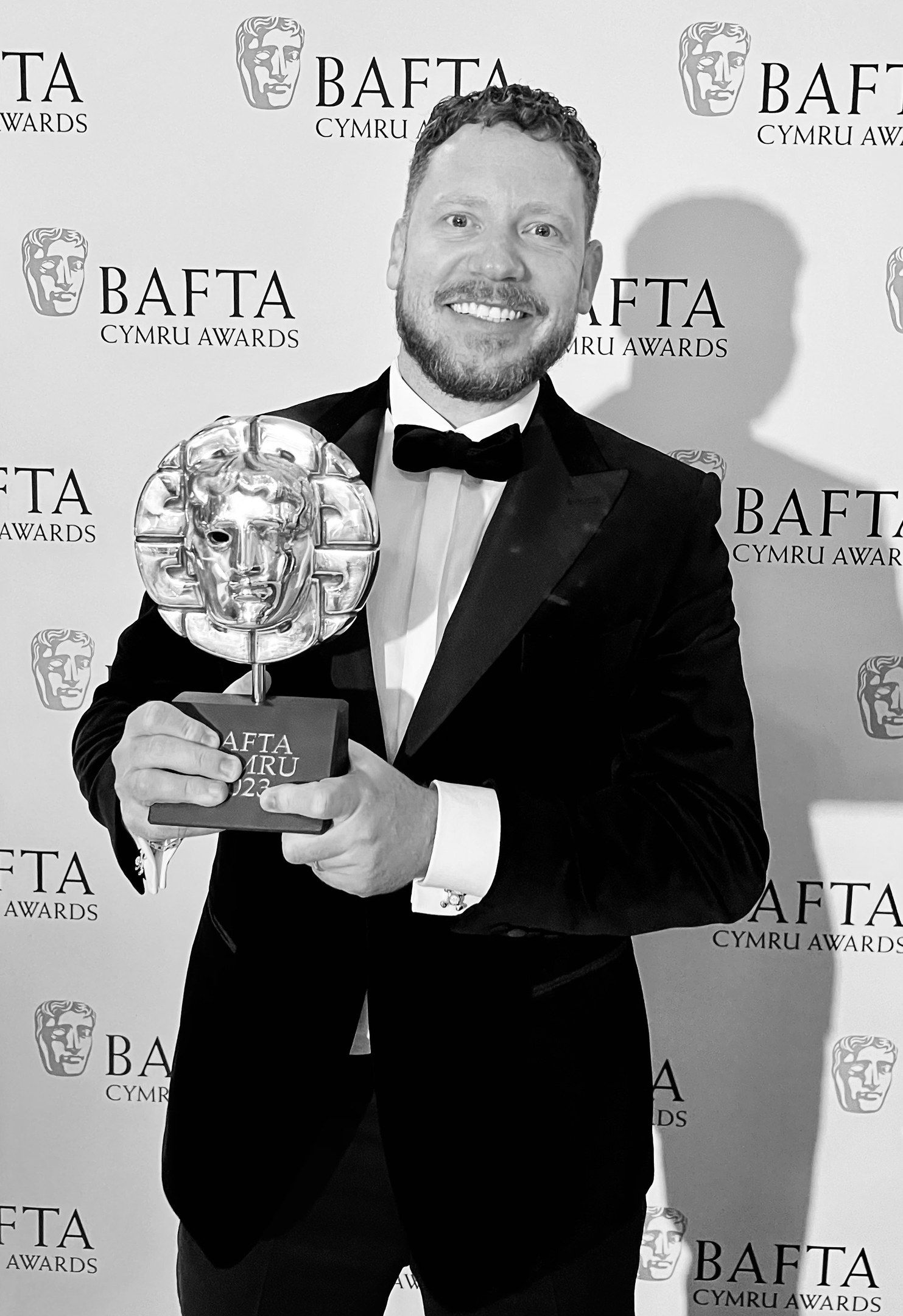 The image shows a man in a tuxedo standing and smiling while holding a BAFTA award in his hands. In...