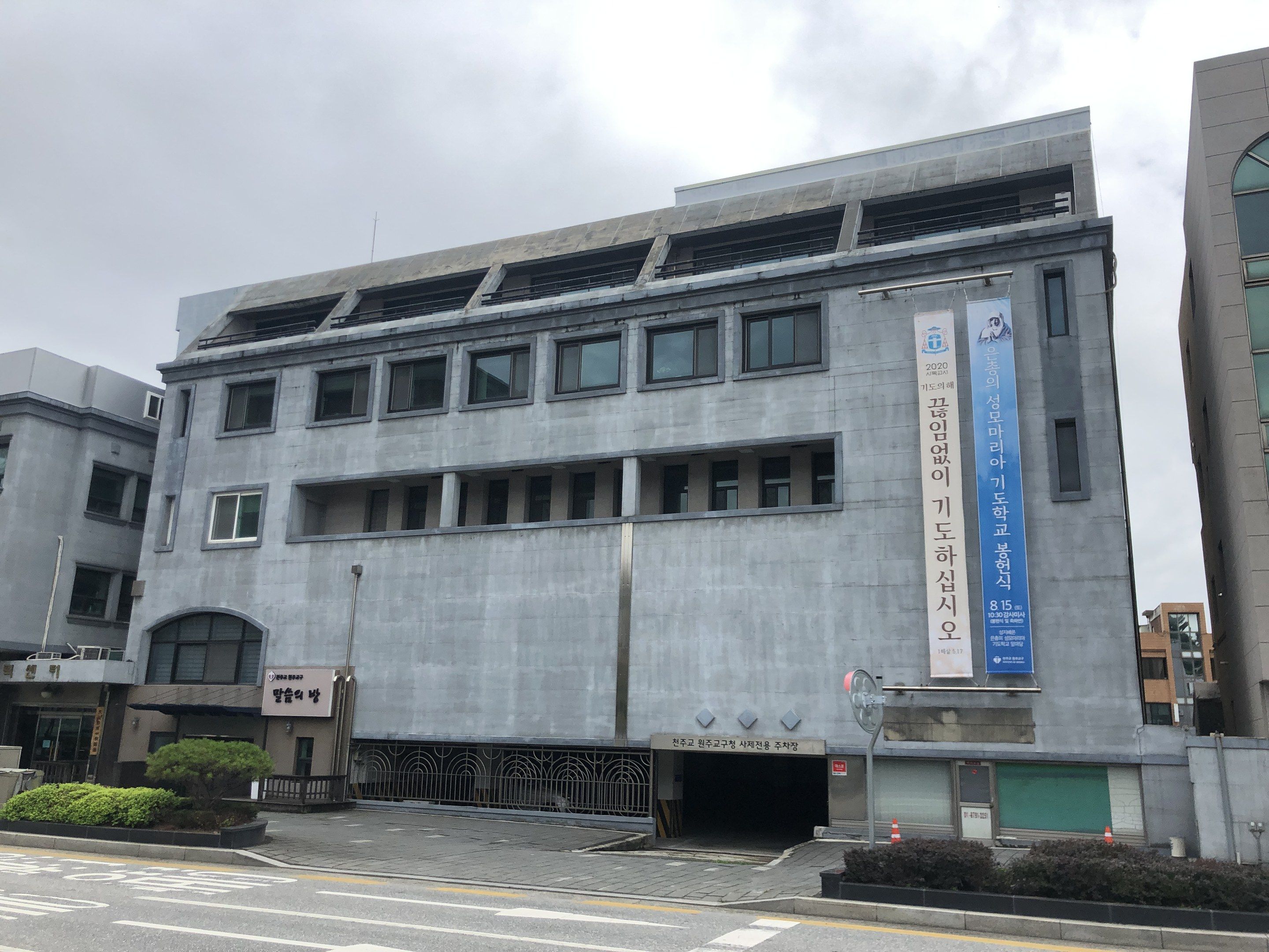 The image shows a large building with a lot of windows on the side of it, surrounded by plants, a...