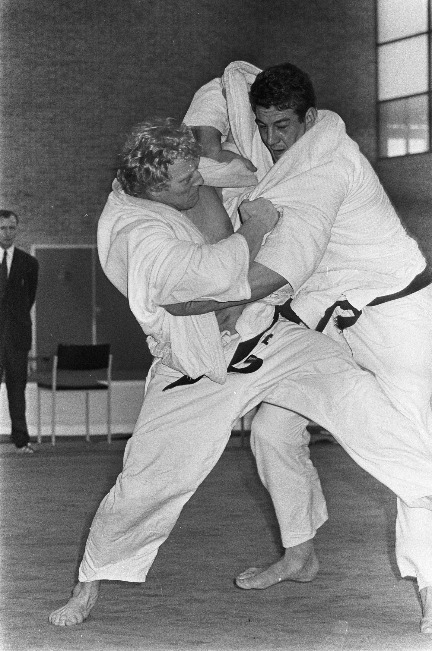 The image shows two men in judo uniforms engaged in a fight in a gym, with a person standing in the...