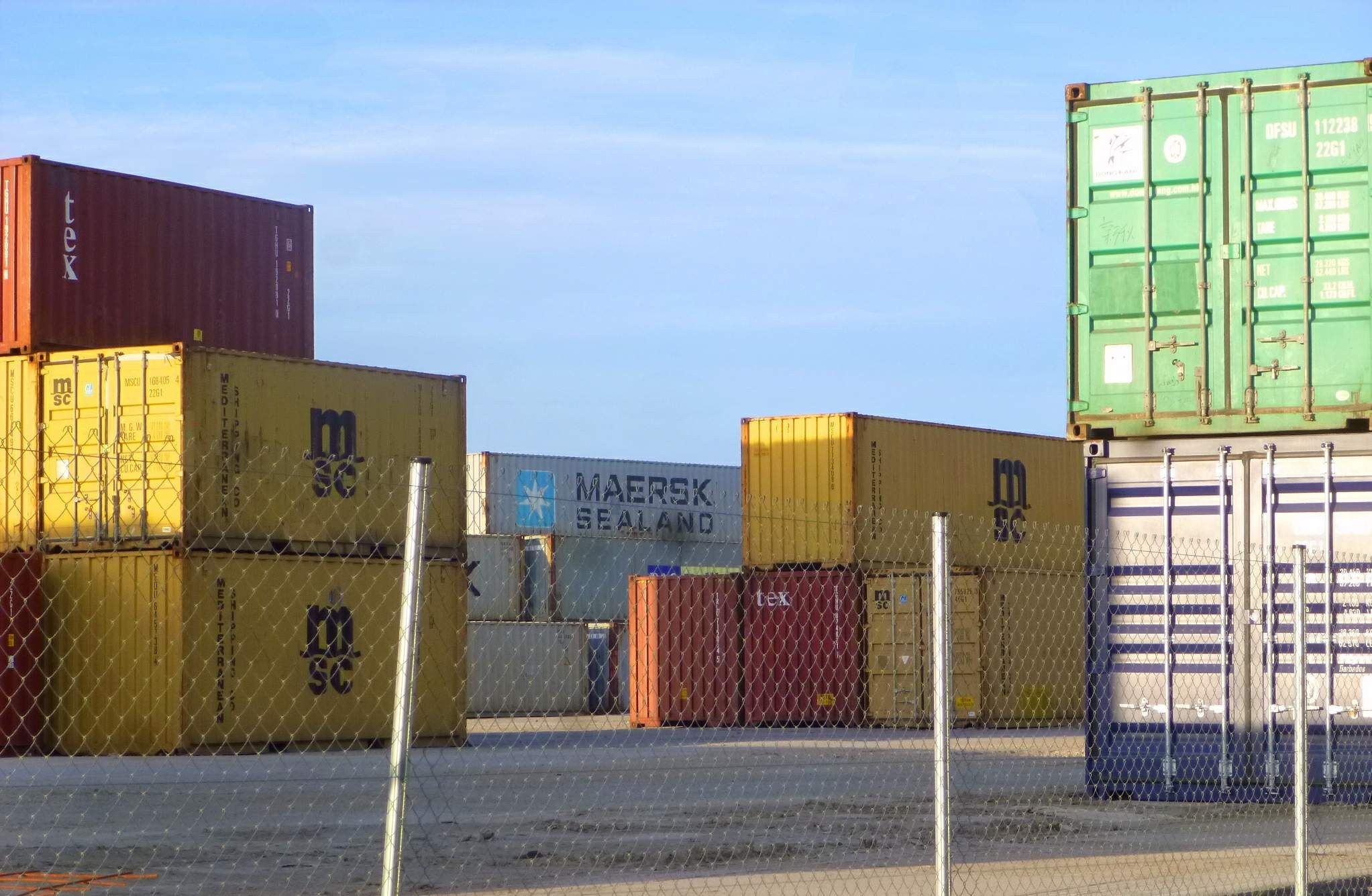 The image shows a large number of shipping containers stacked on top of each other, with a mesh...