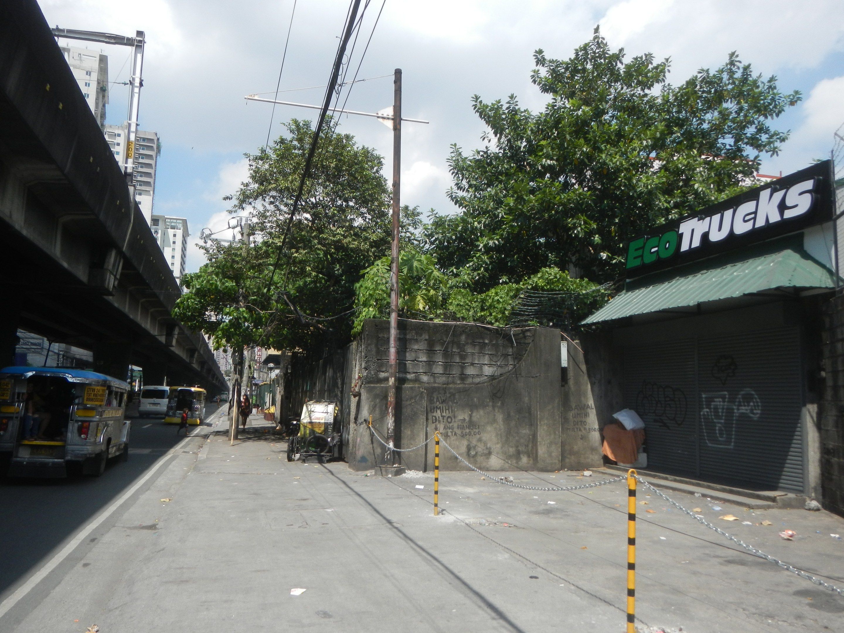 The image shows a city street with vehicles driving on it, a bridge on the left side, buildings in...