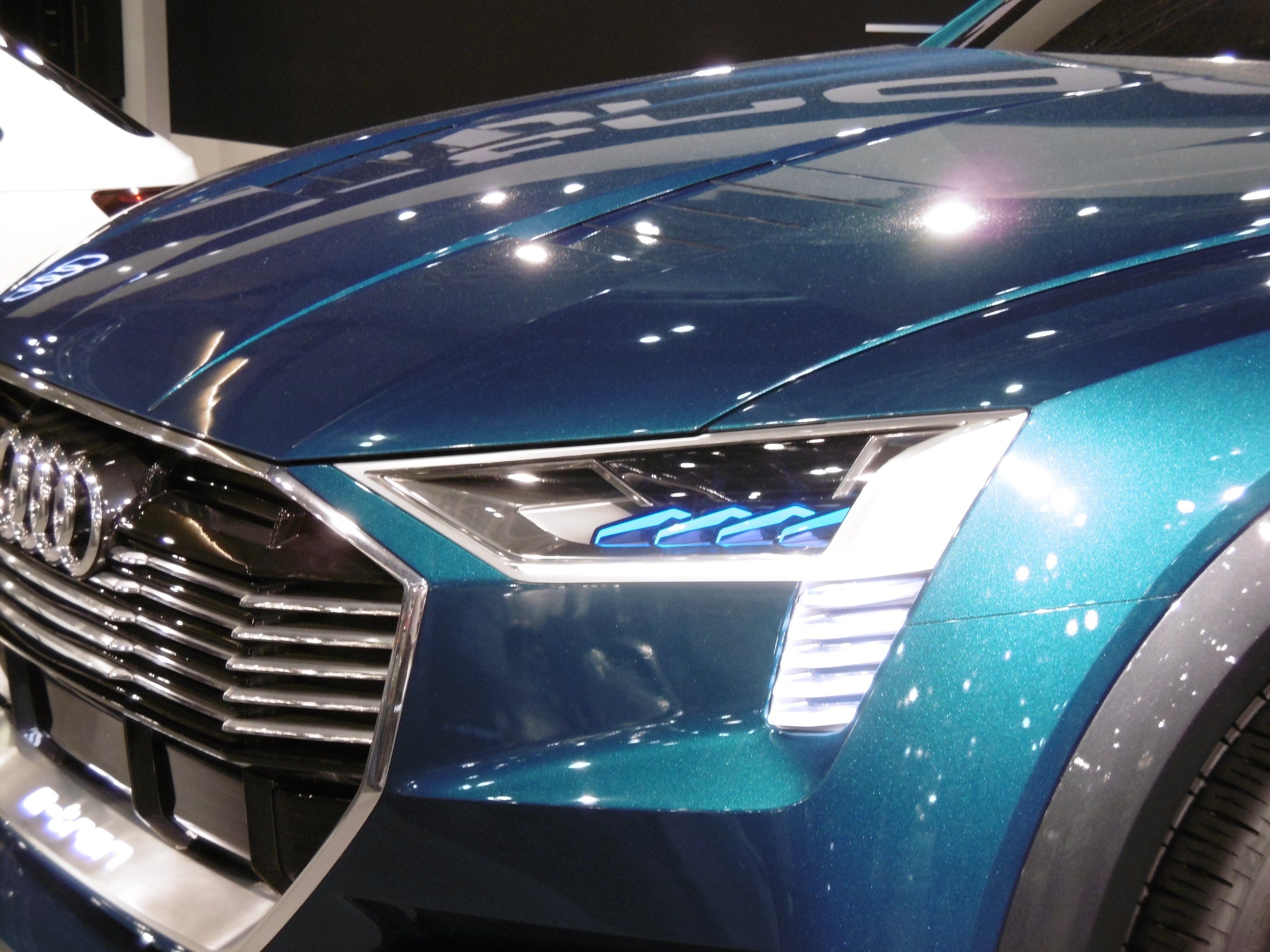 The image shows a blue Audi e-tron concept car on display at an auto show, with a white car in the...