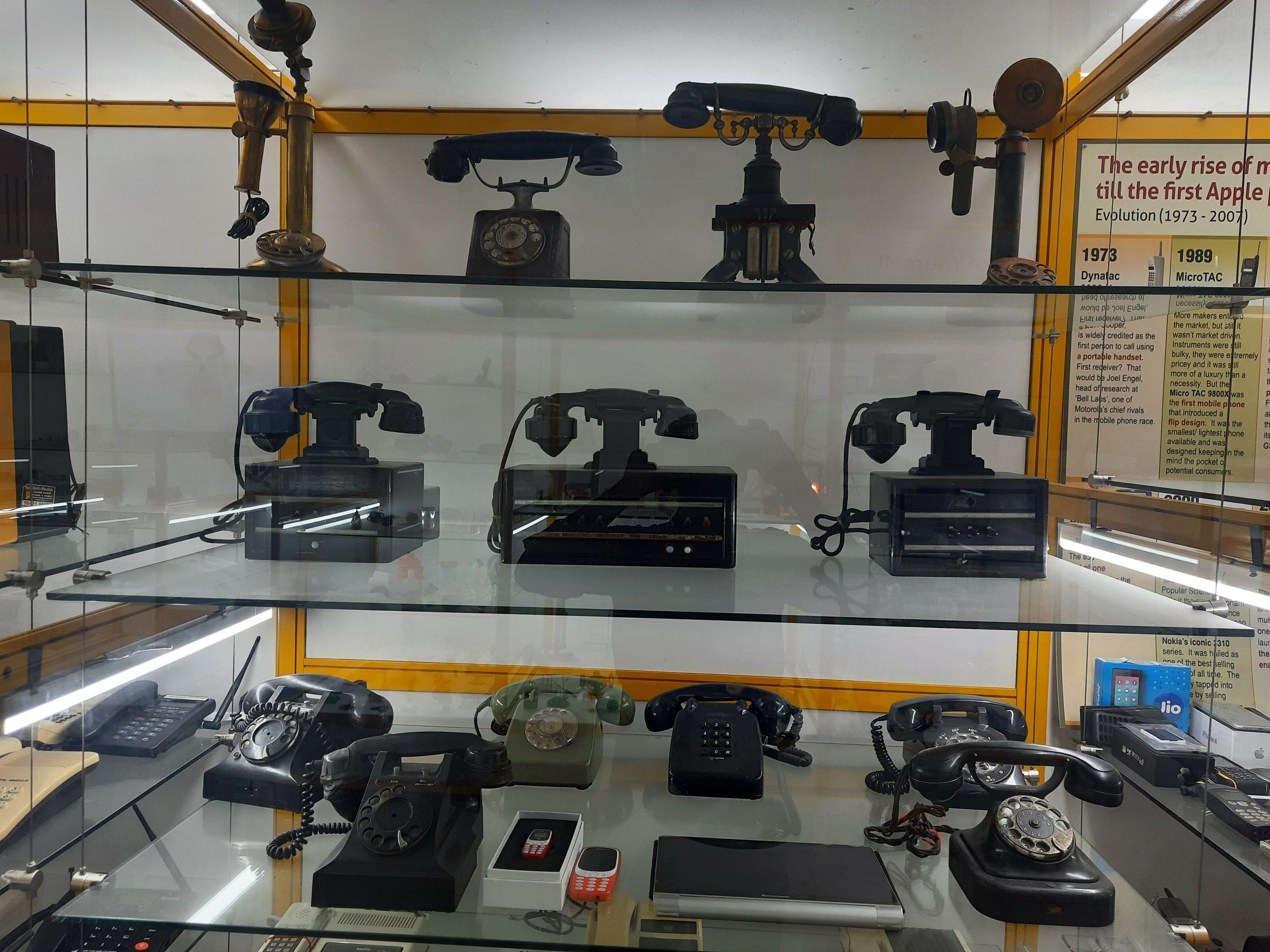 The image shows a display case filled with lots of different types of telephones on glass shelves,...