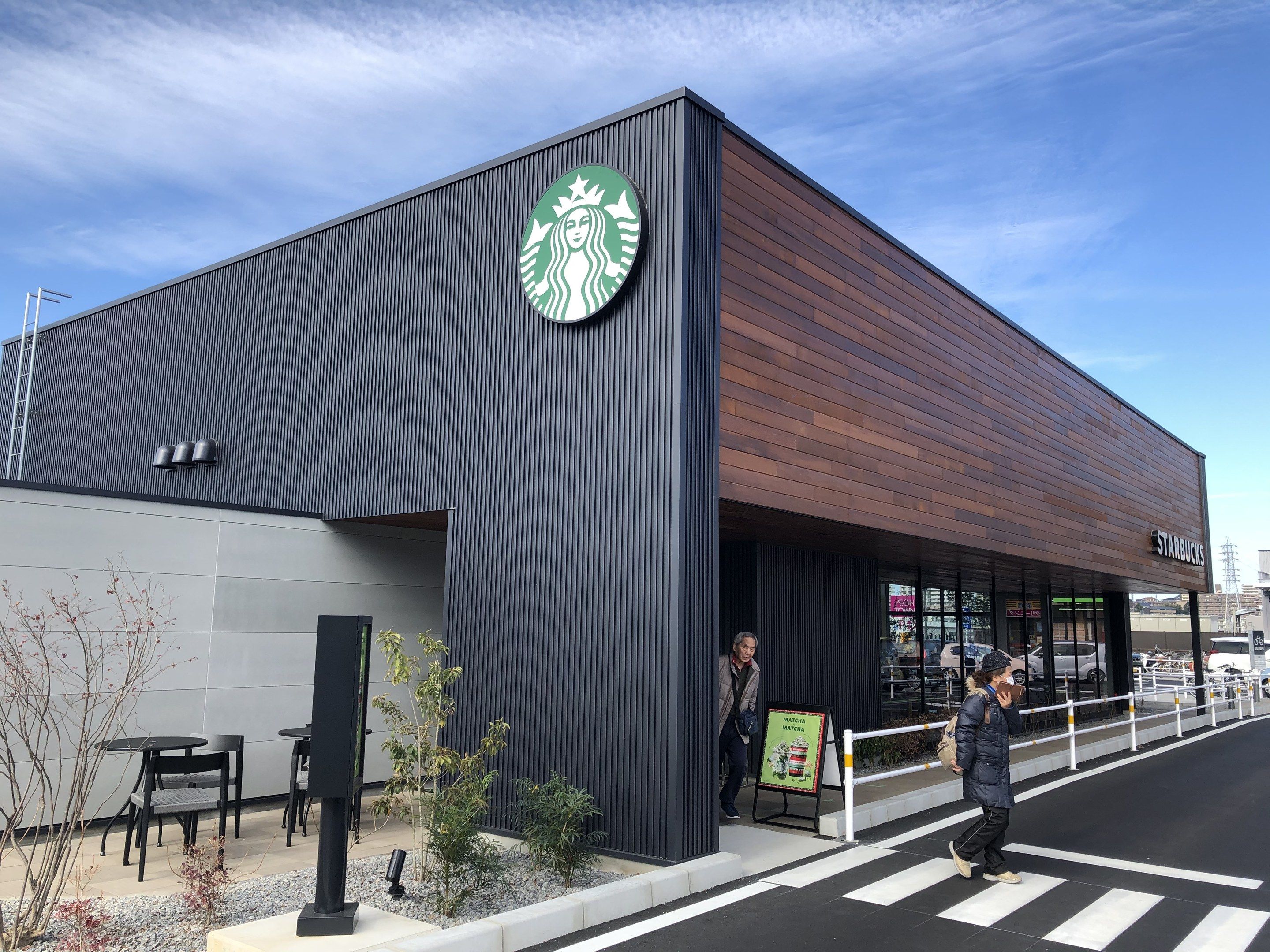 The image shows a Starbucks coffee shop on the corner of a street, with two people walking on the...