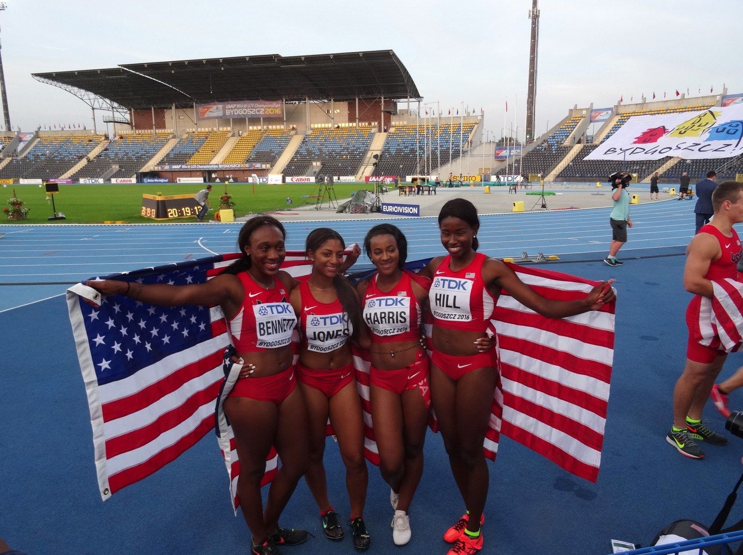 The image shows a group of four women standing next to each other on a track, smiling and holding...