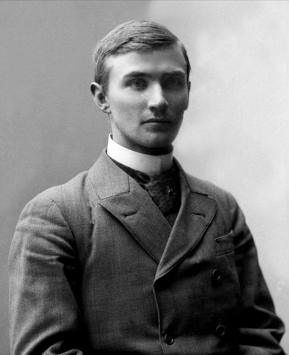 The image shows a black and white photo of a man in a suit and tie, believed to be the founder of...