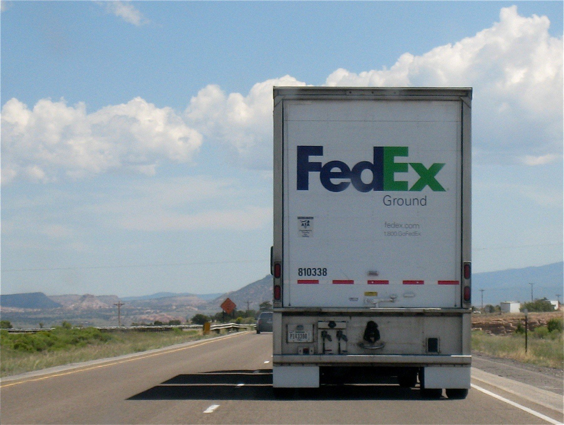 The image shows a FedEx truck driving down a highway with mountains in the background. The truck is...
