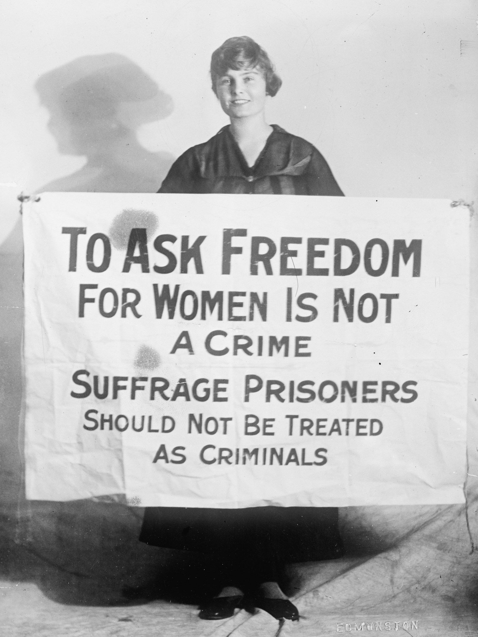 The image shows a woman standing and smiling while holding a banner that reads "To Ask Freedom for...