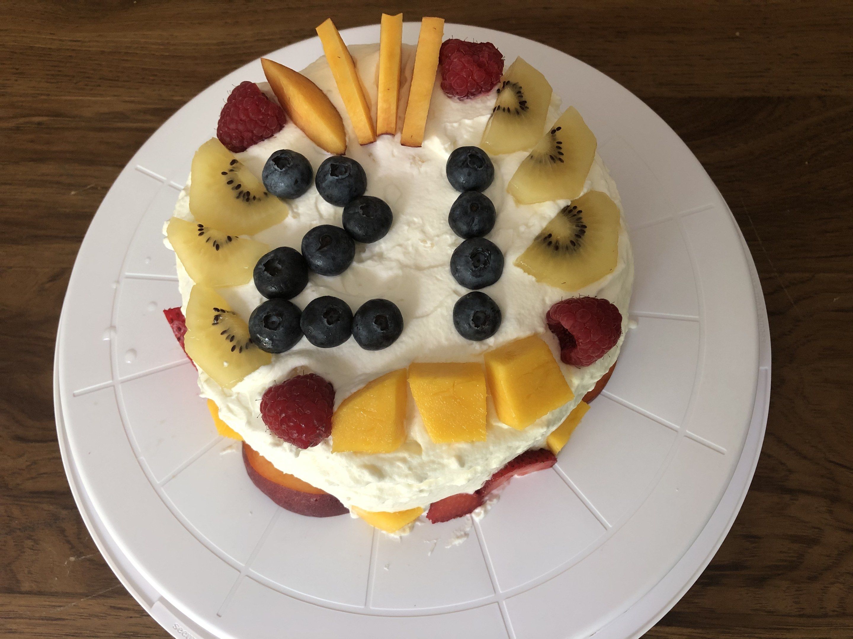 The image shows a white plate on a wooden table with a cake decorated with various fruits arranged...