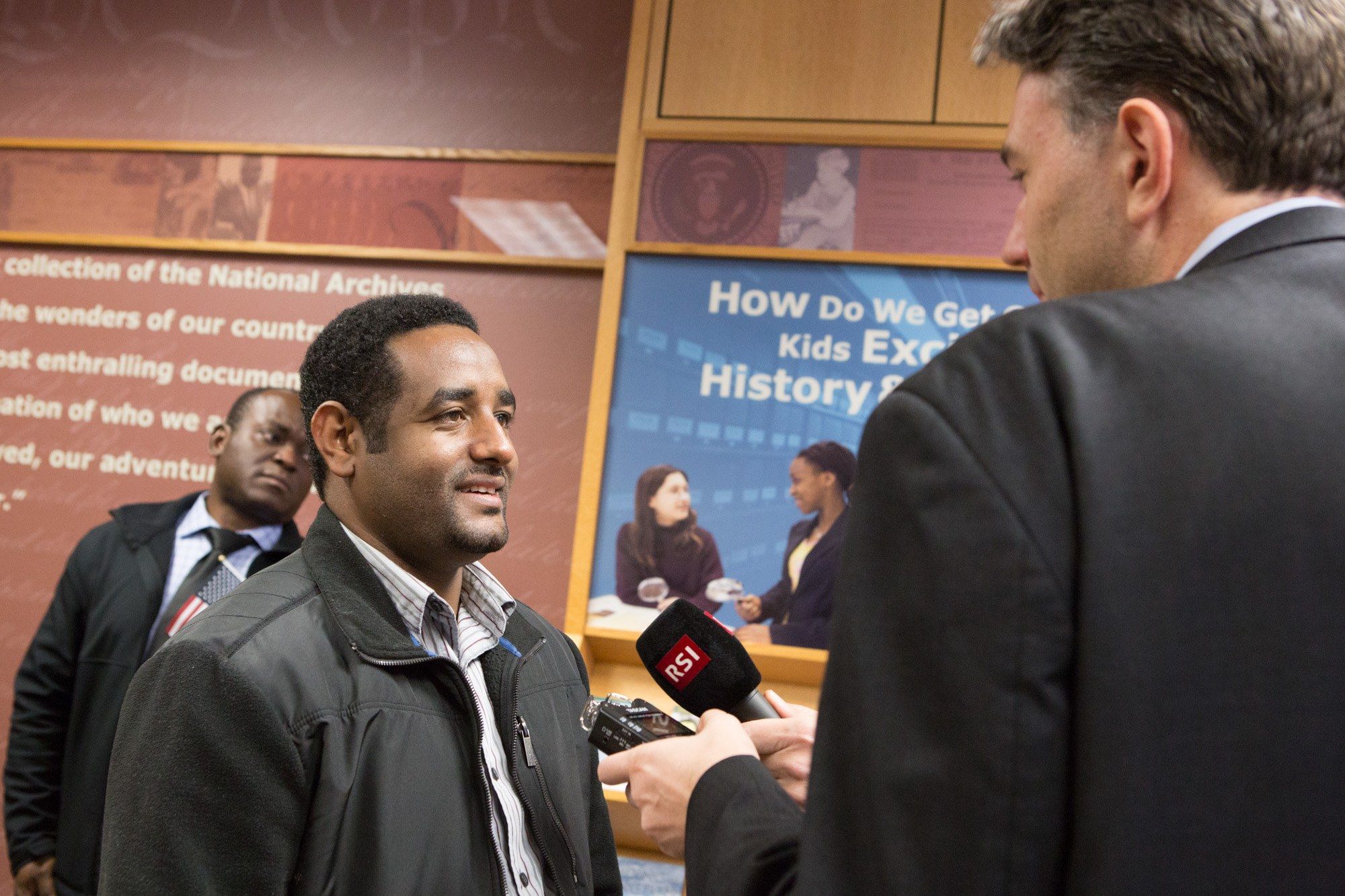 The image shows a man in a suit talking to a reporter in front of a wall adorned with banners. The...