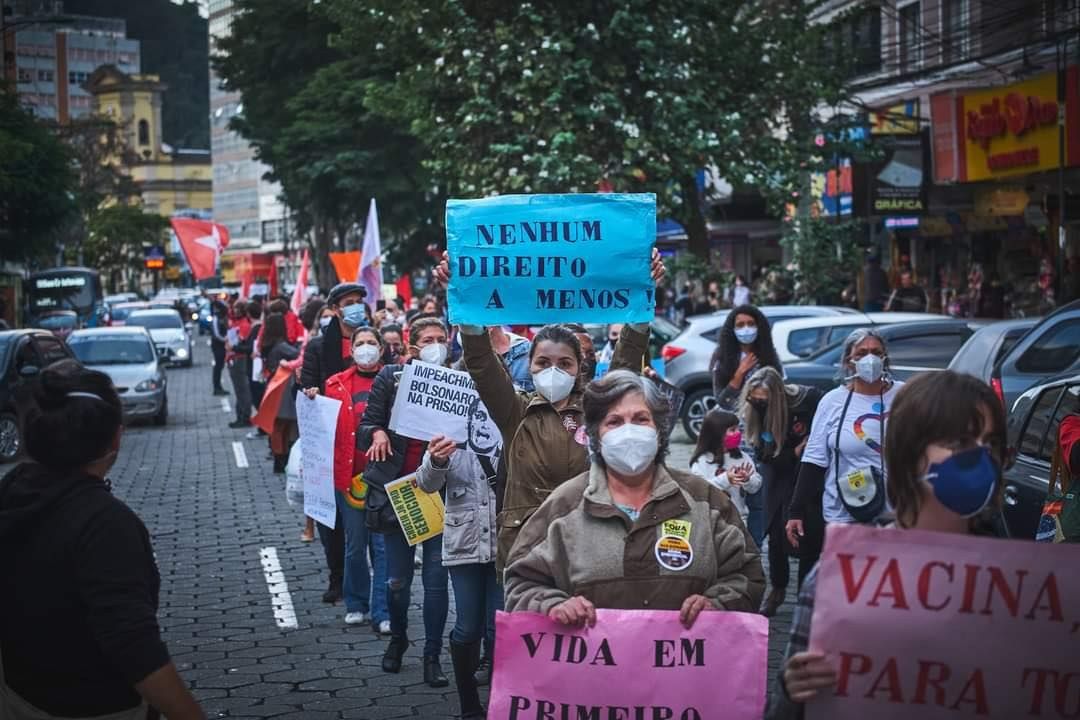 The image shows a group of people wearing masks and holding signs in the middle of a street,...