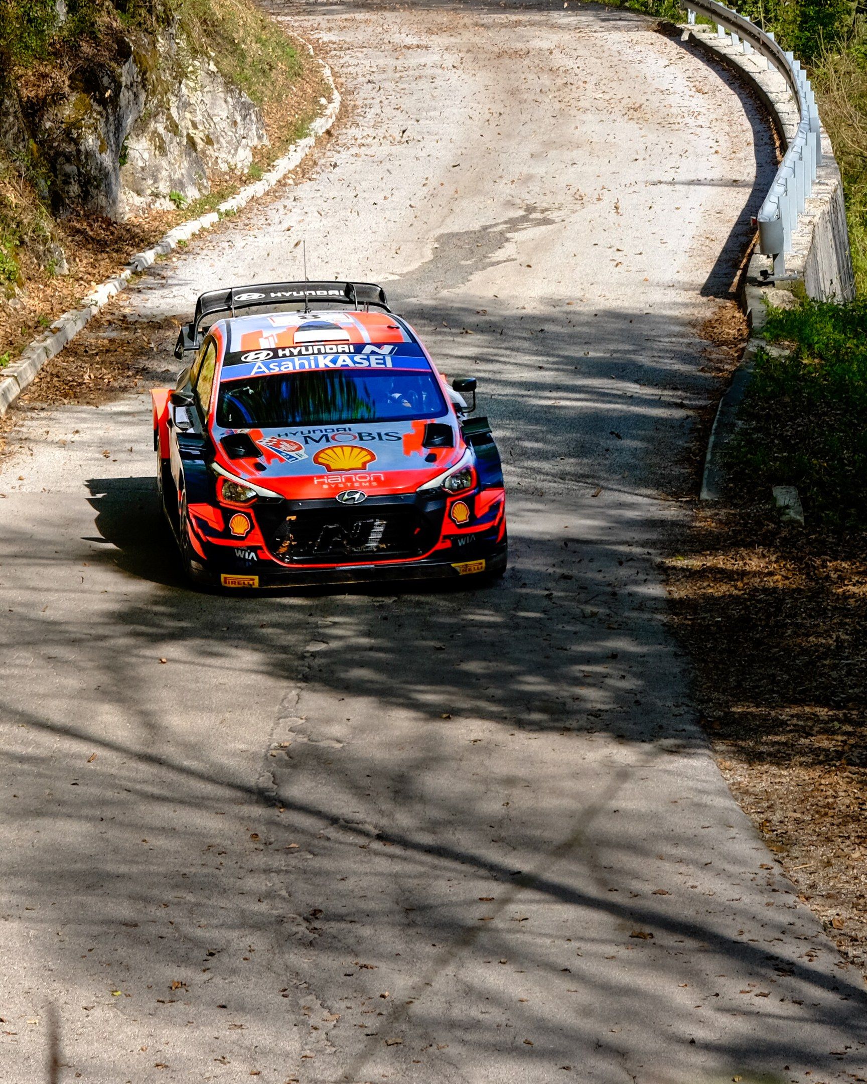 The image shows a rally car driving down a winding road surrounded by trees and plants on either...