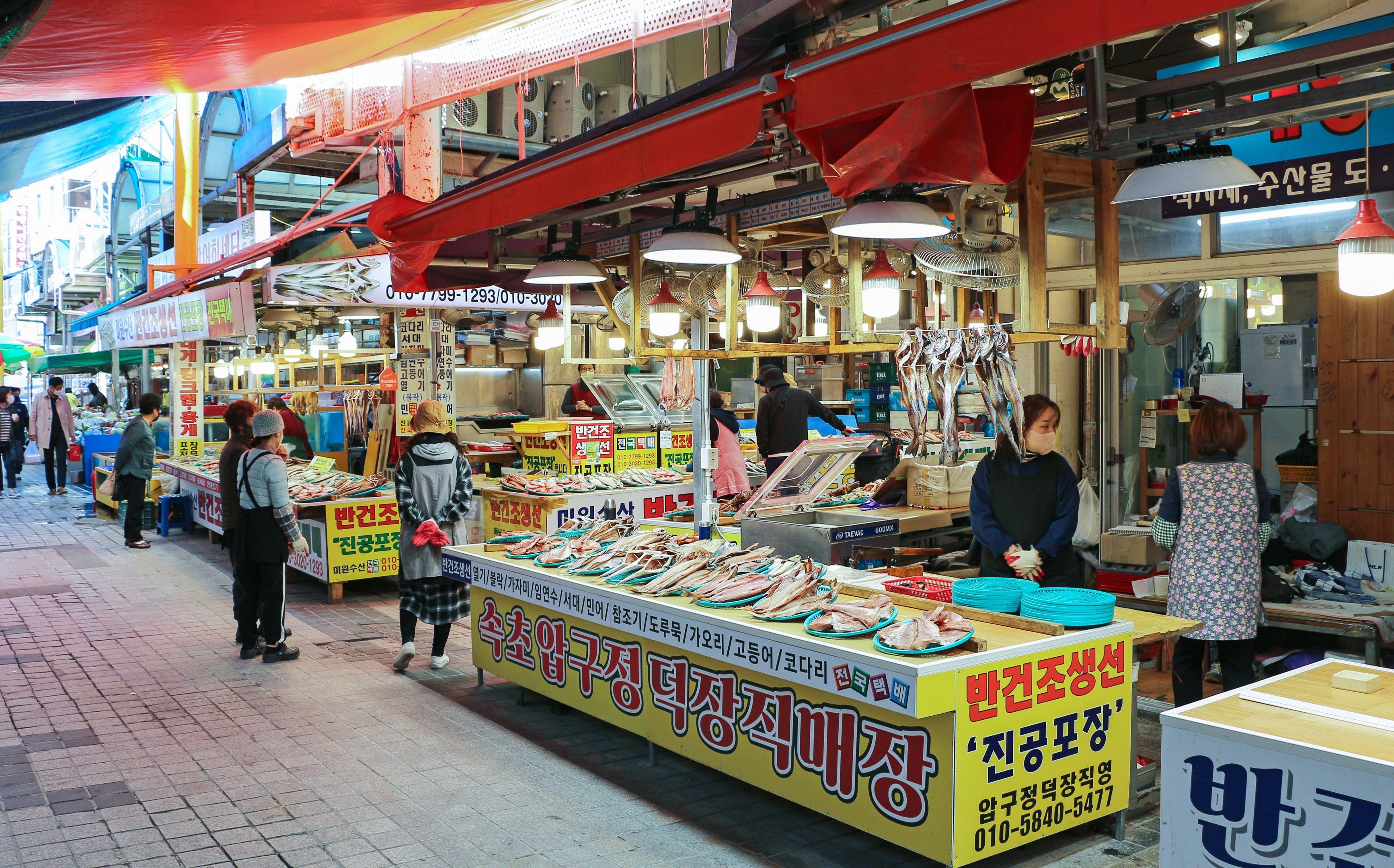 The image shows a bustling street market in Seoul, South Korea. There are many people walking...