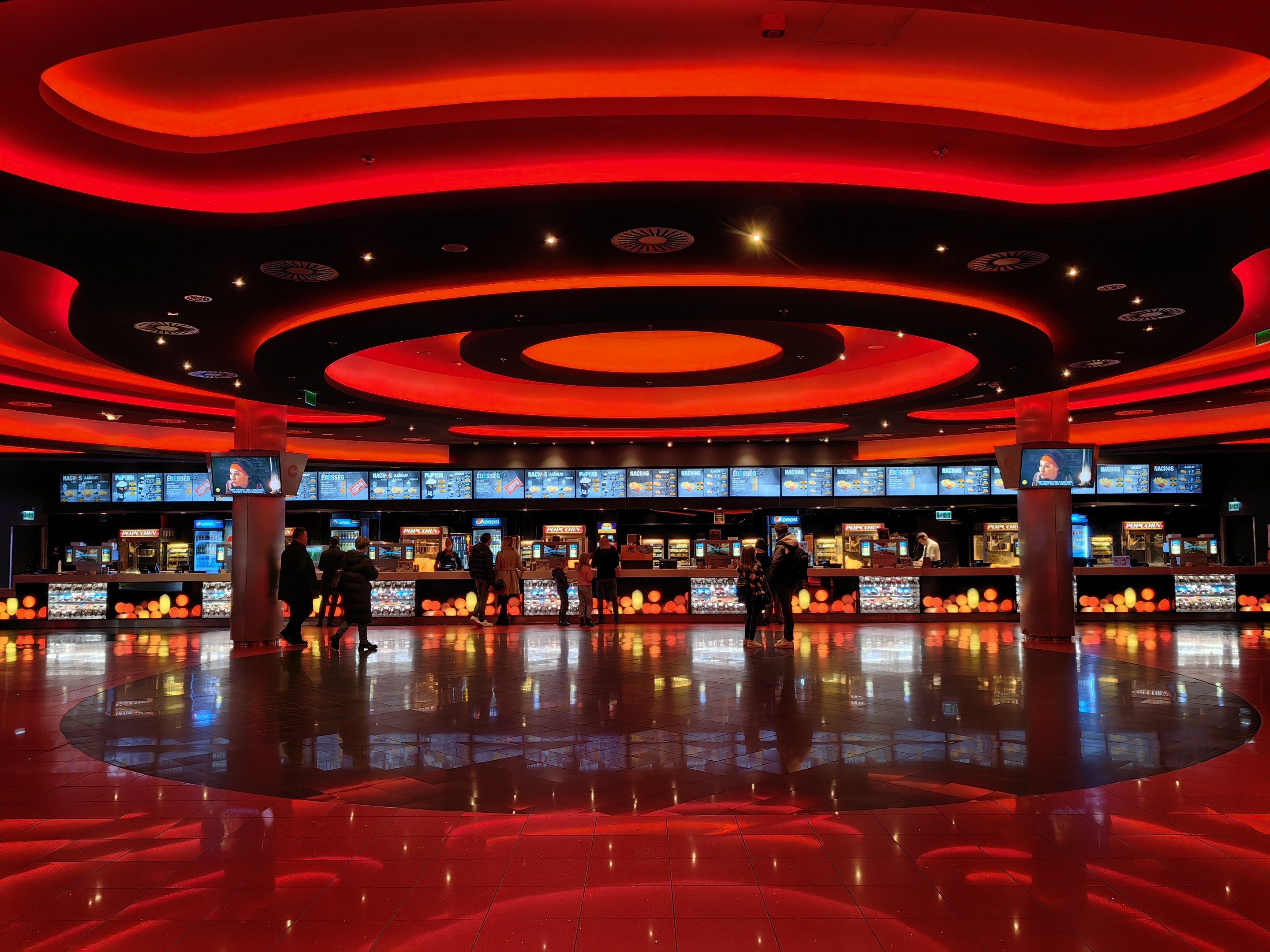 The image shows a large room filled with lots of people, some standing and some walking on the...