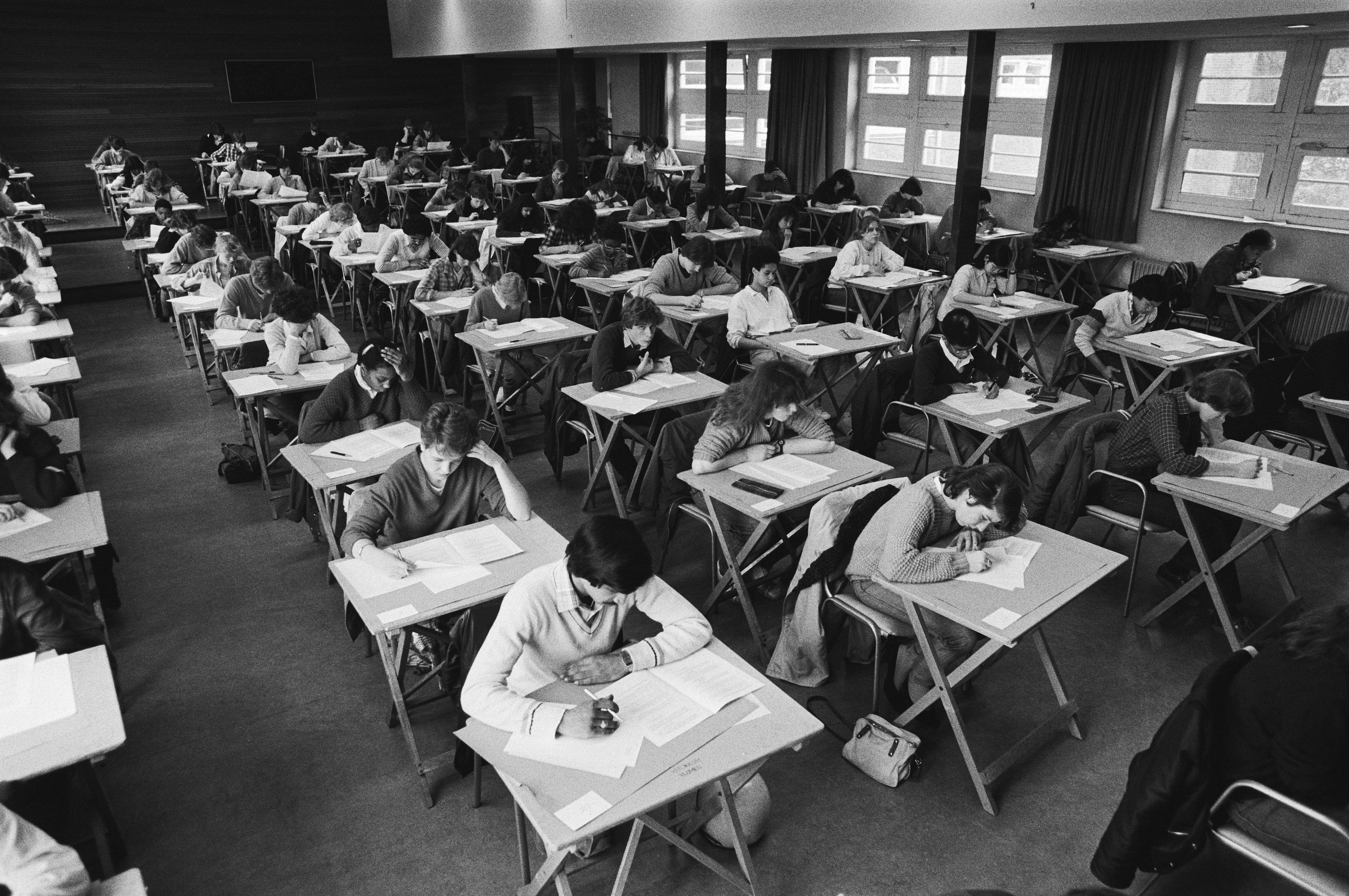 The image shows a black and white photo of a classroom full of students sitting at desks, writing...
