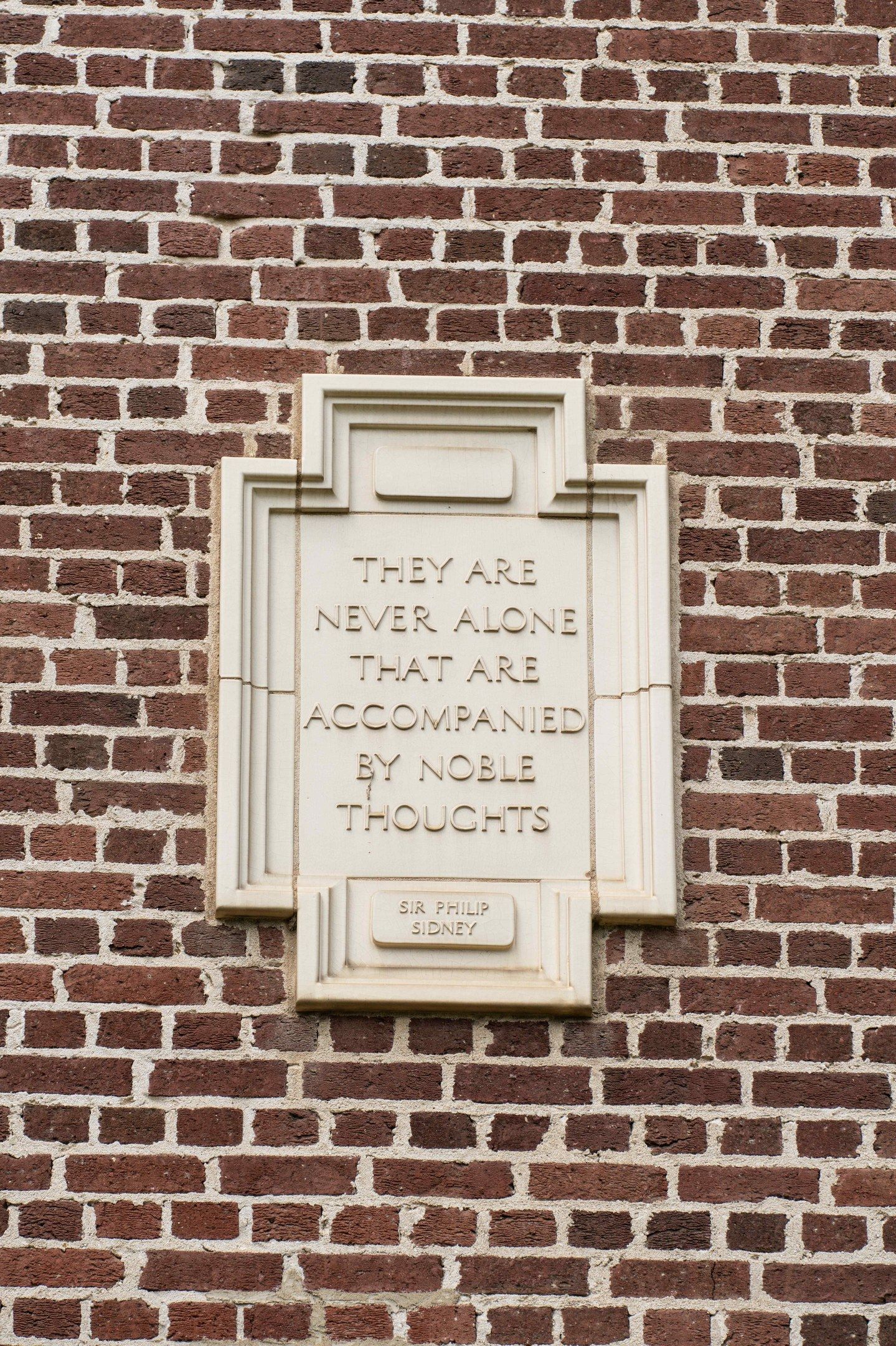 The image shows a brick wall with a plaque on it that reads "They are never alone that are...