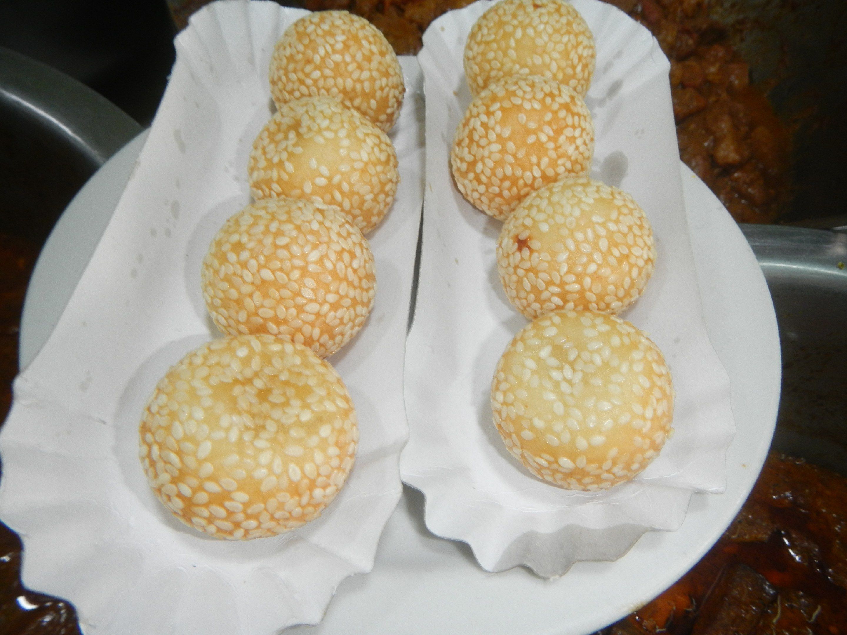 The image shows a white plate topped with six sesame buns on top of a table, with a bowl of food in...