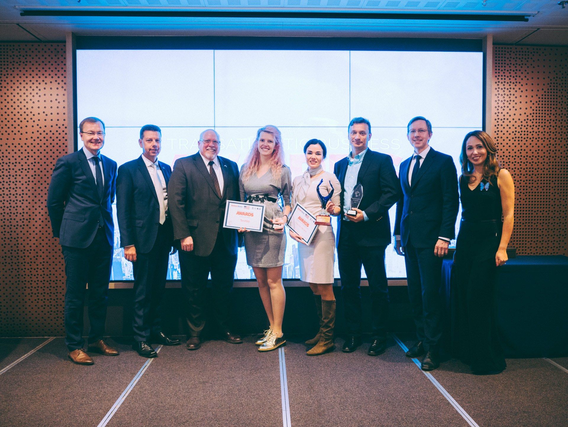 The image shows a group of people standing next to each other in front of a screen, smiling and...