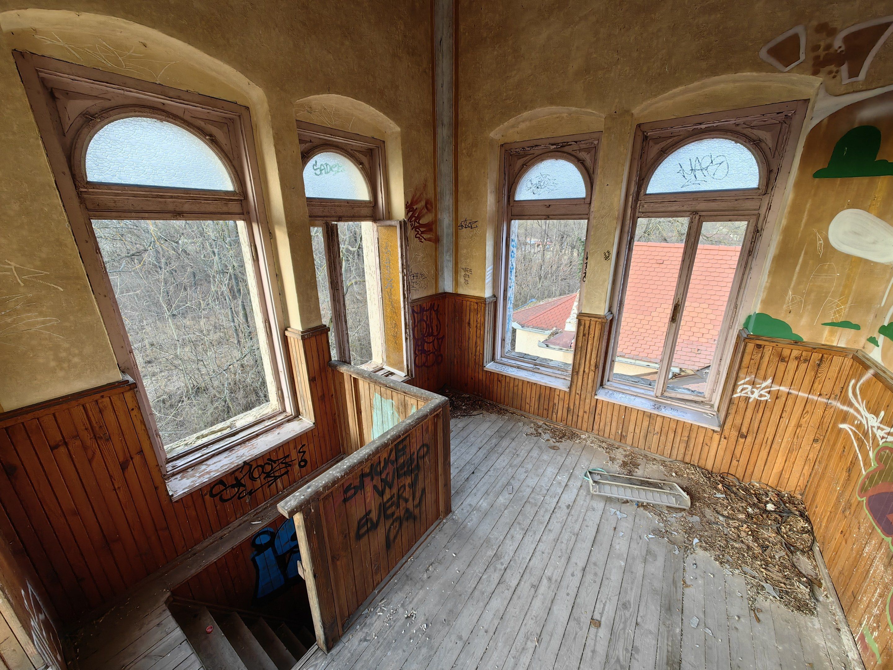 The image shows an abandoned building with graffiti on the walls and windows, a wooden floor, a...