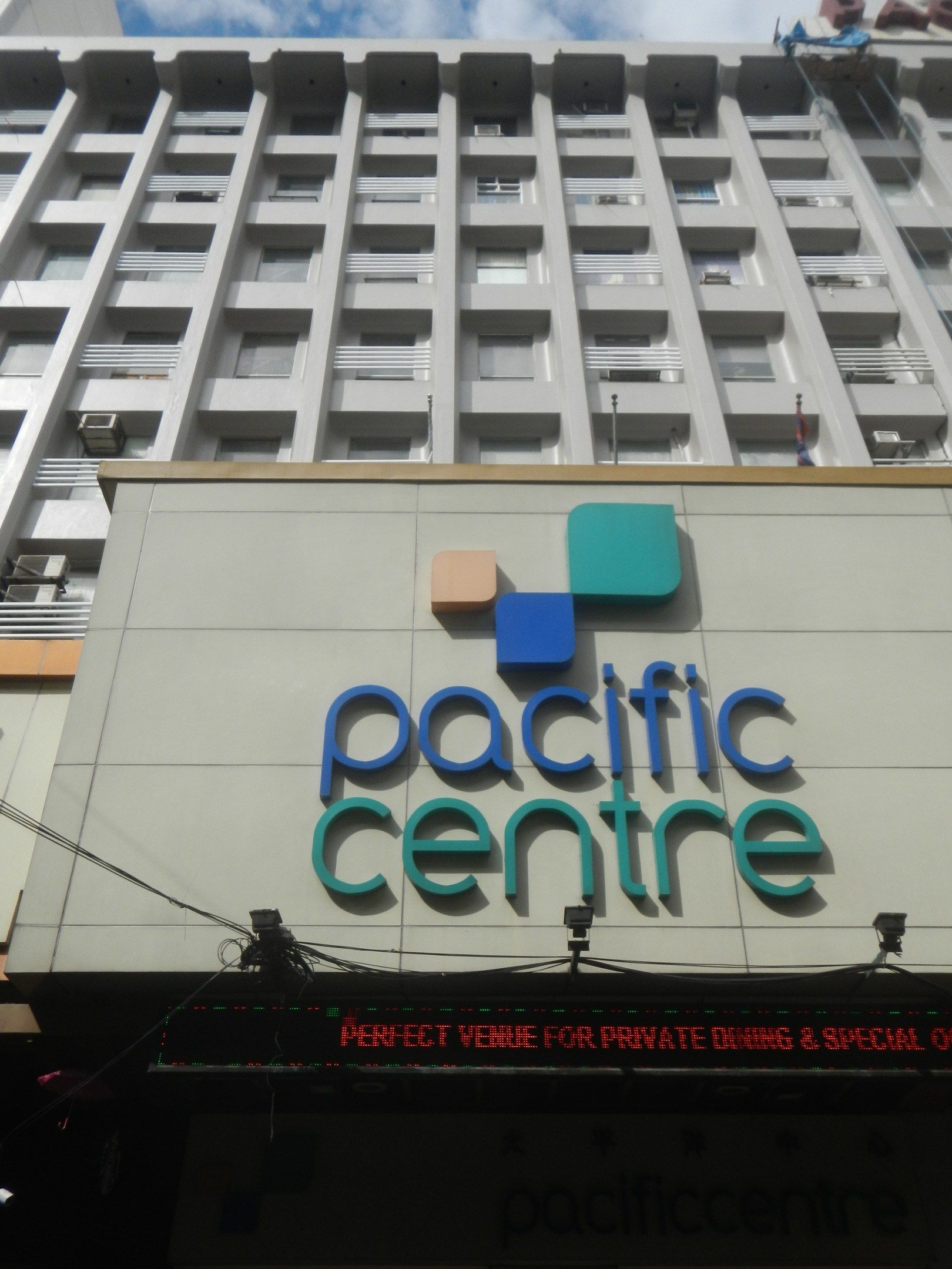 The image shows the Pacific Centre Hotel in Bangkok, Thailand. It is a building with windows, air...