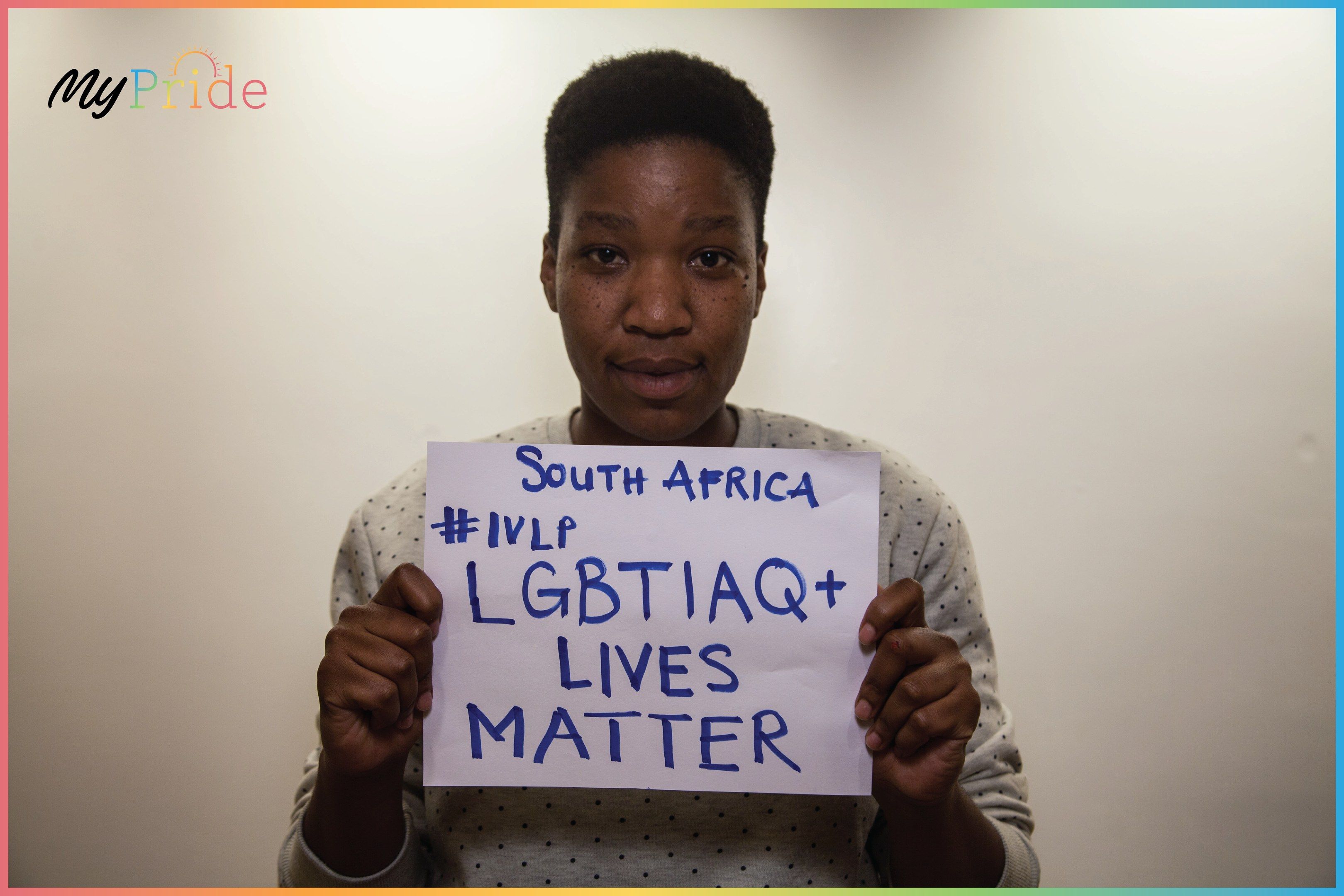 The image shows a woman standing in front of a wall, holding up a sign that reads "LGBTQ+ Lives...