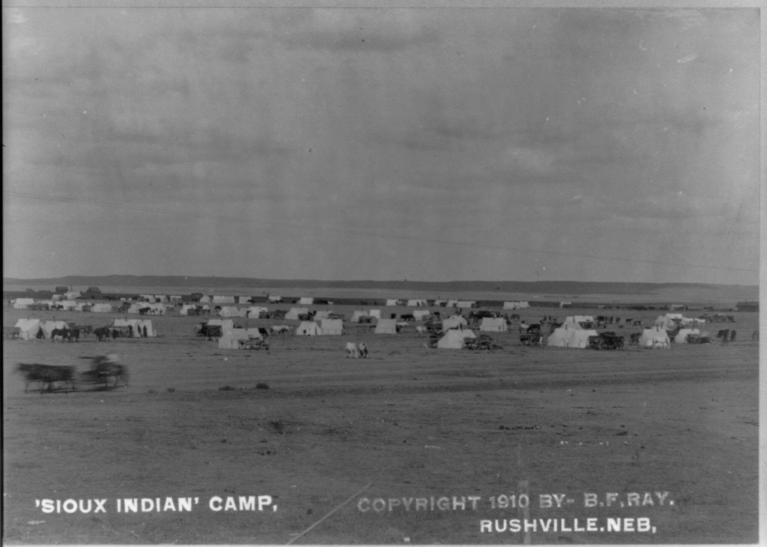 The image shows a black and white photo of a Sioux Indian camp in Rushville, Nebraska. There are...