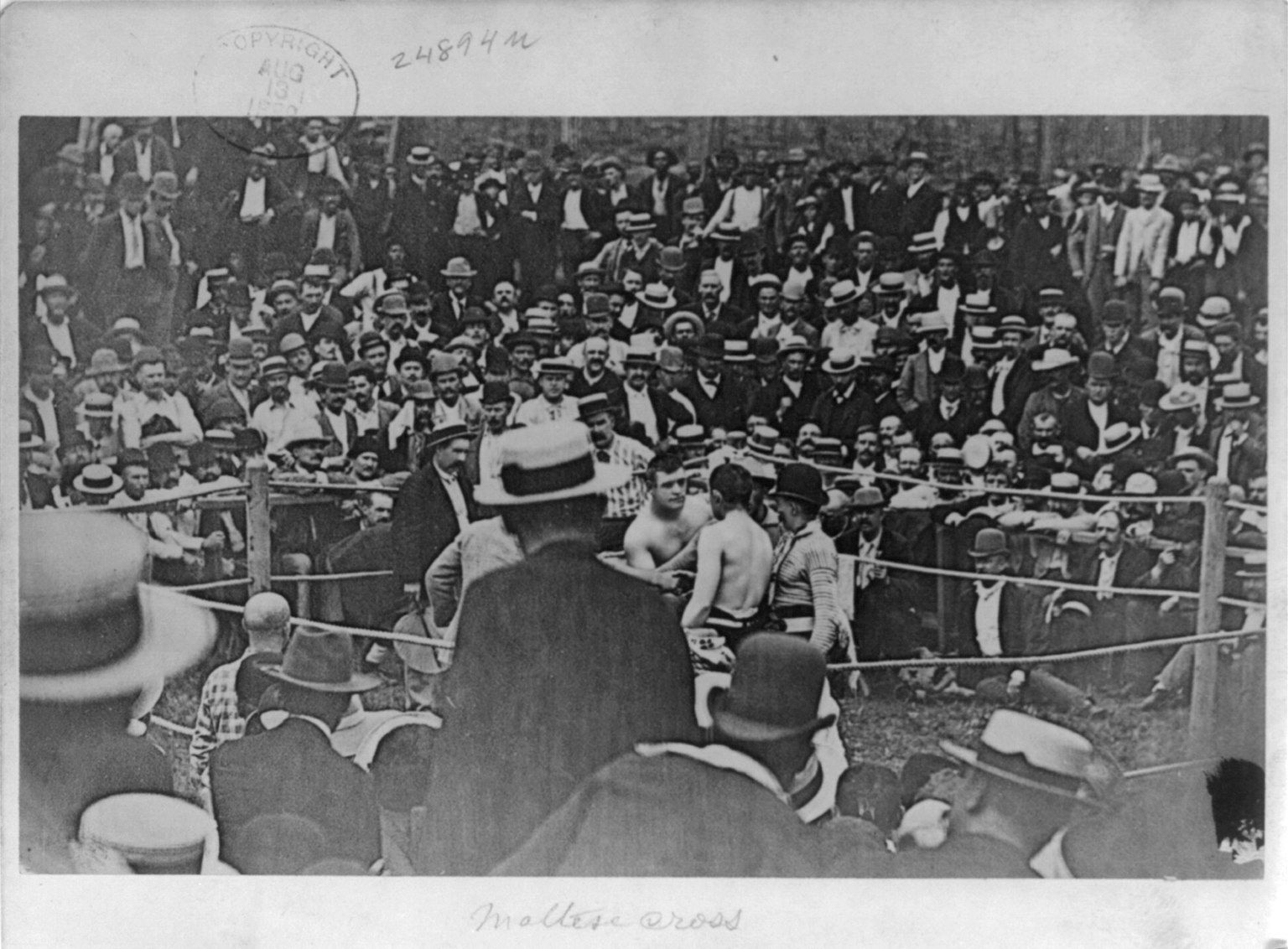 The image shows a black and white photo of a crowd of people in a boxing ring, with some of them...