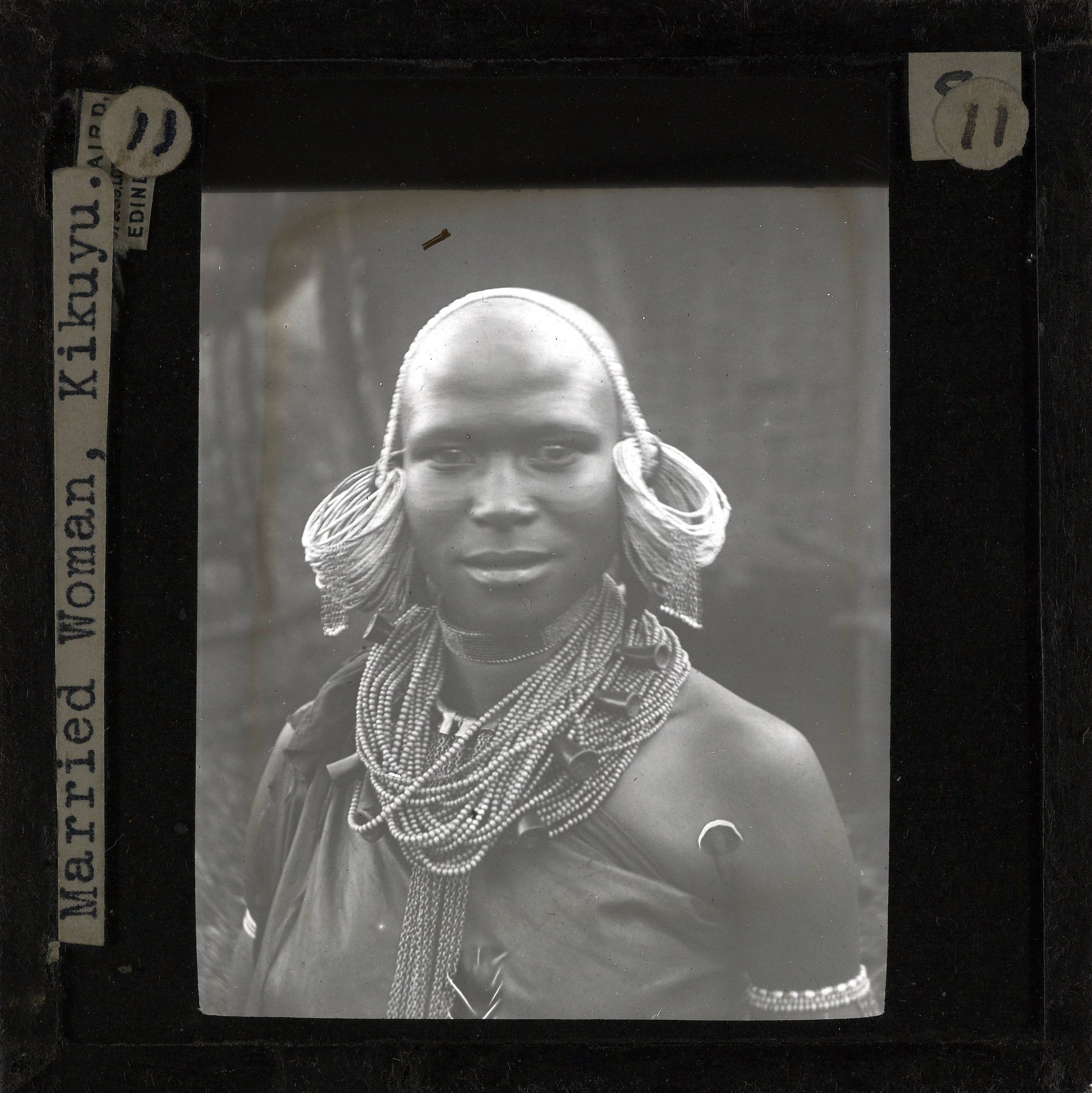 The image shows a black and white photo of a woman from the Kikuyu tribe wearing ornaments. The...